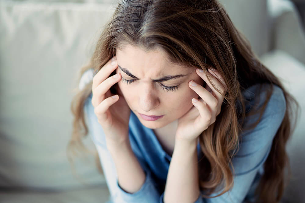 A young woman sits on a sofa holding her temples with a pained expression, suggesting headache or migraine discomfort