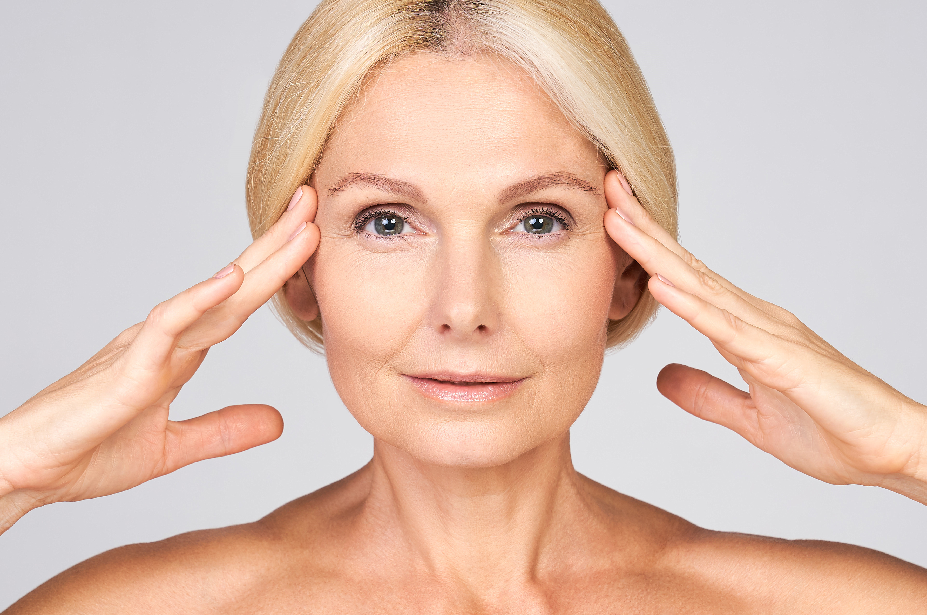 A mature woman with blonde hair touches her temples while showcasing smooth, youthful facial skin against a plain gray backgr