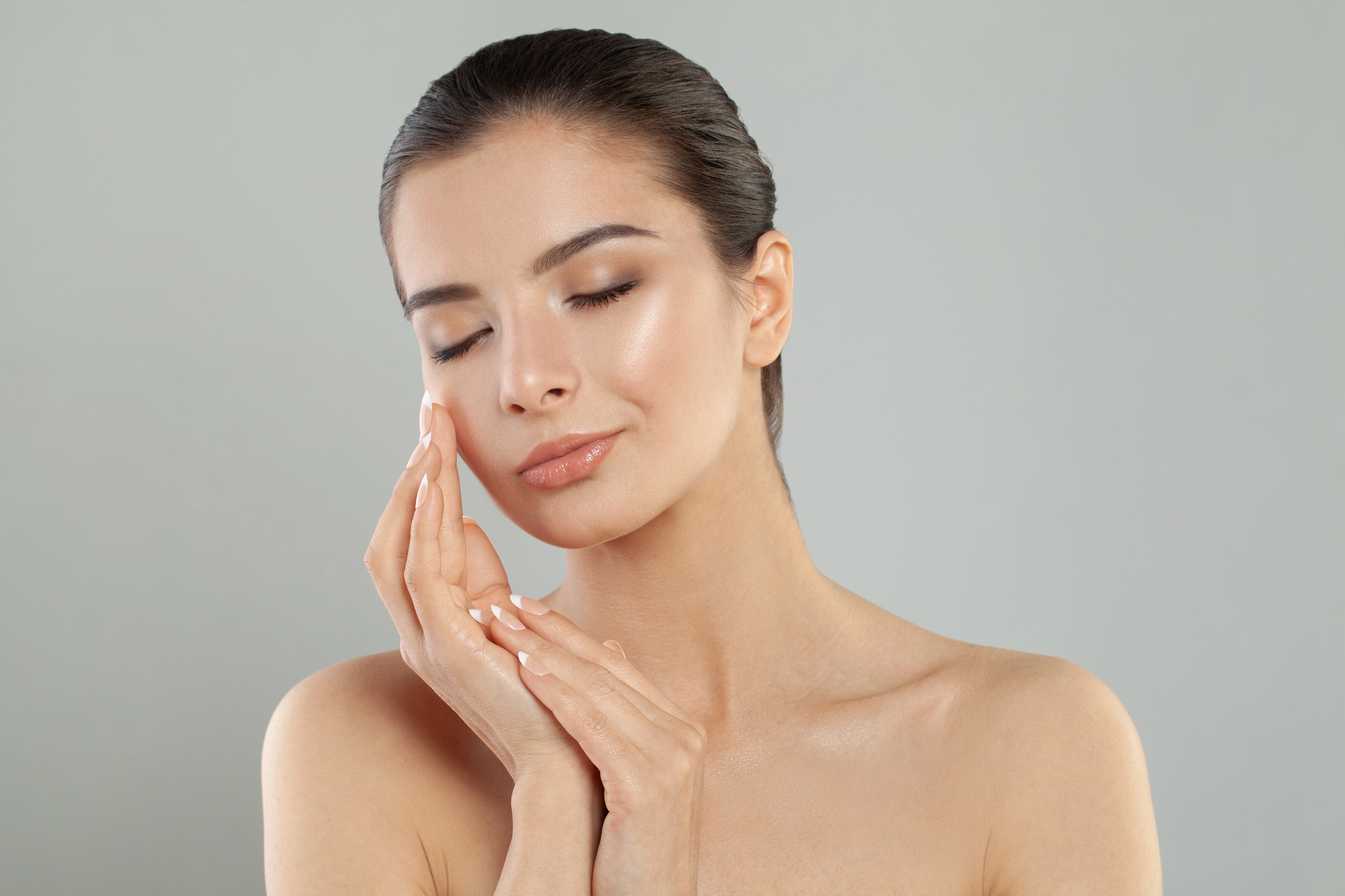 A woman with sleek hair and bare shoulders gently touches her glowing, flawless skin against a neutral gray background