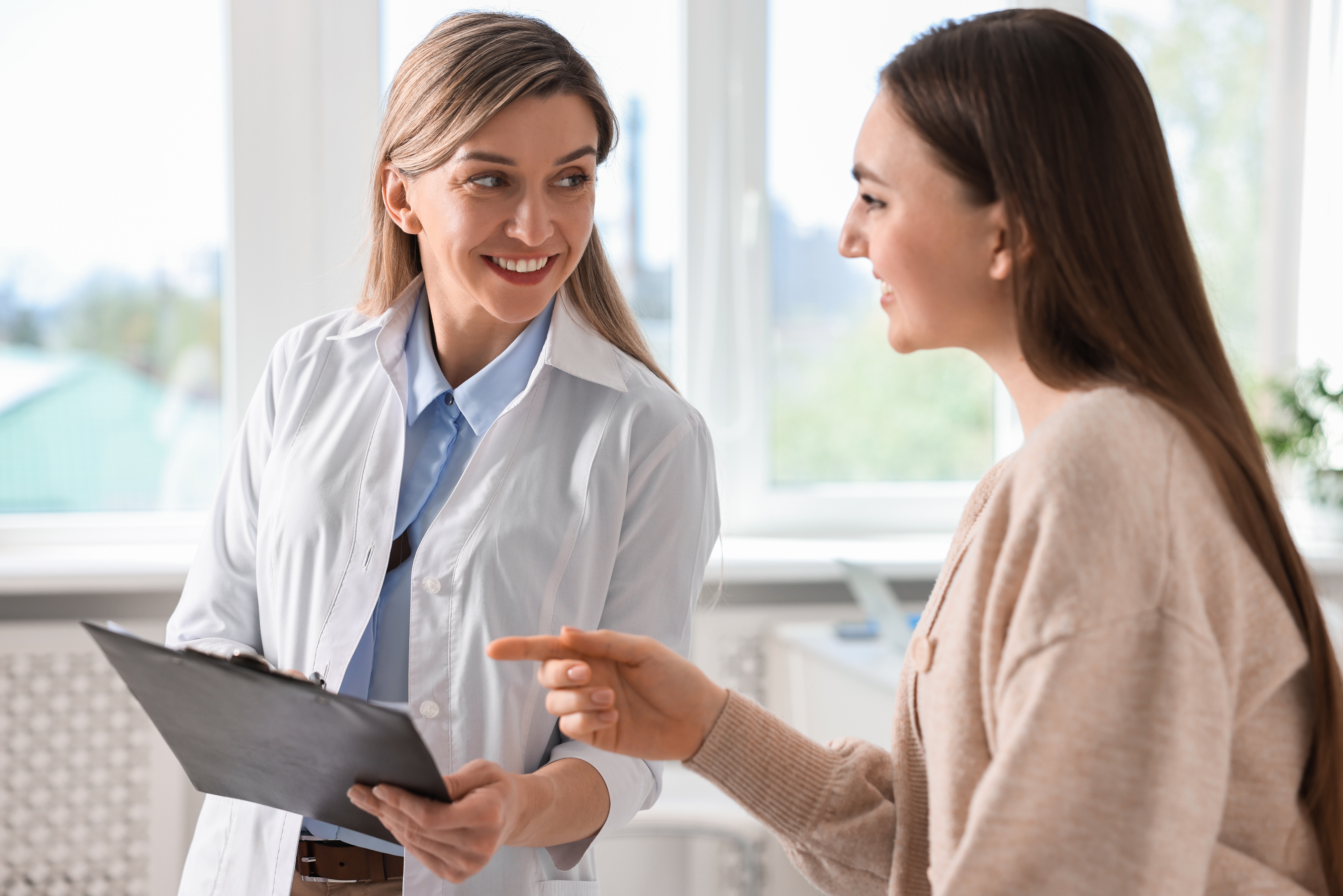 A smiling female doctor in a white coat holds a clipboard while discussing treatment with a female patient in a bright office.