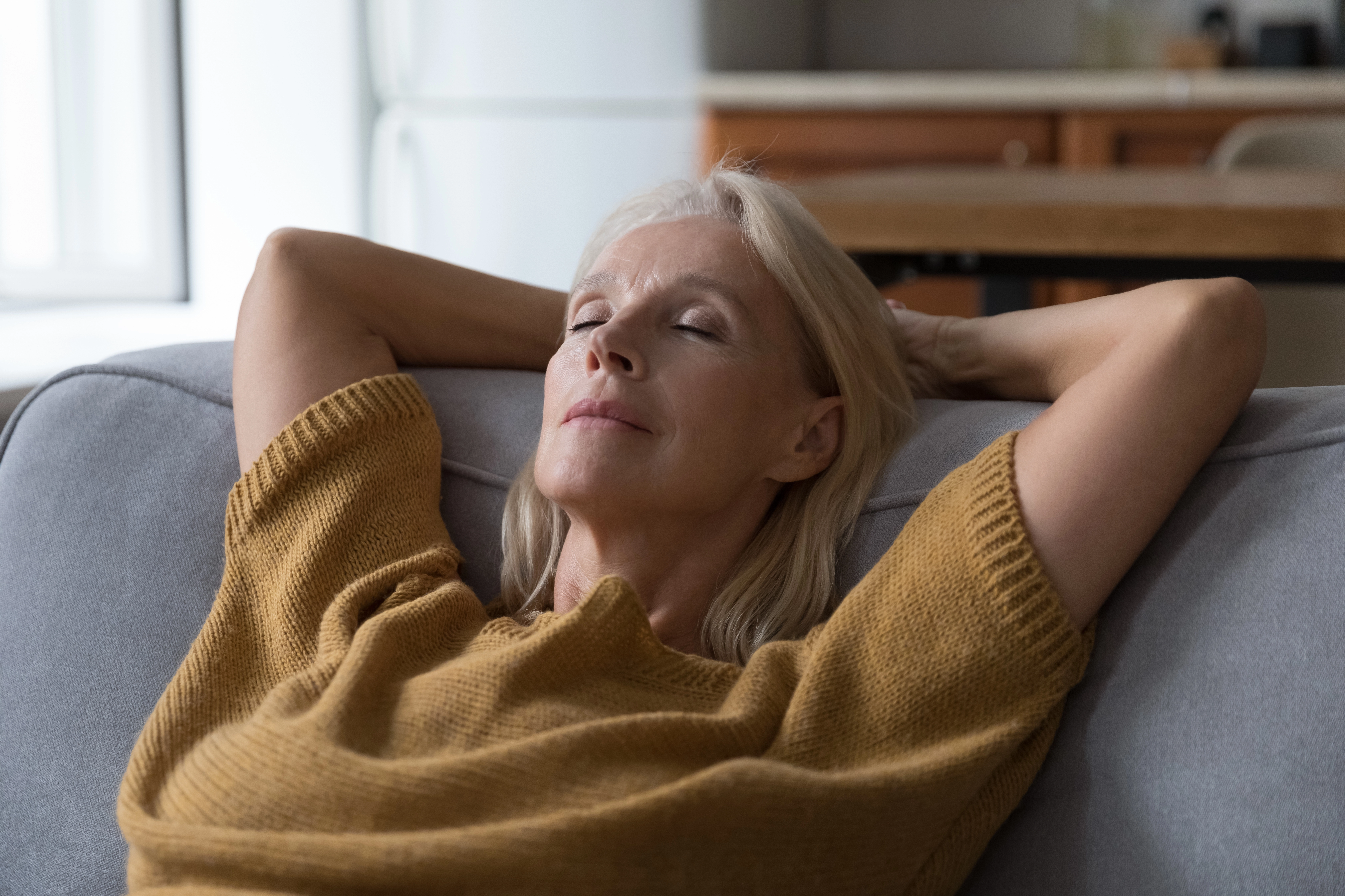 A relaxed older woman reclines on a sofa with her eyes closed and hands behind her head, enjoying a peaceful moment