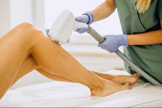 A provider in scrubs and gloves performs laser hair removal on a patient's leg using a handheld device