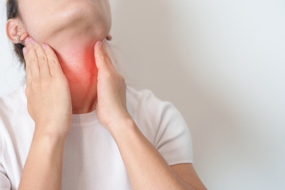A woman touches her neck with both hands, with a red highlighted area indicating throat or thyroid pain