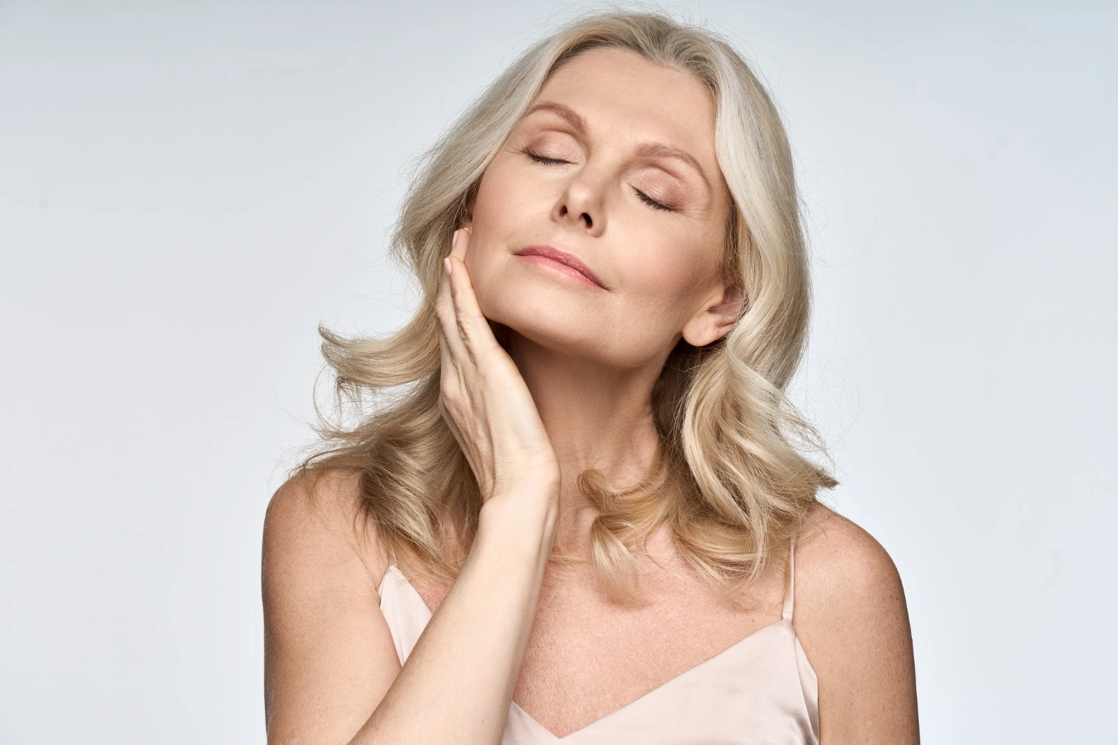 A mature woman with closed eyes gently touches her neck and jawline, showcasing smooth skin against a plain light background