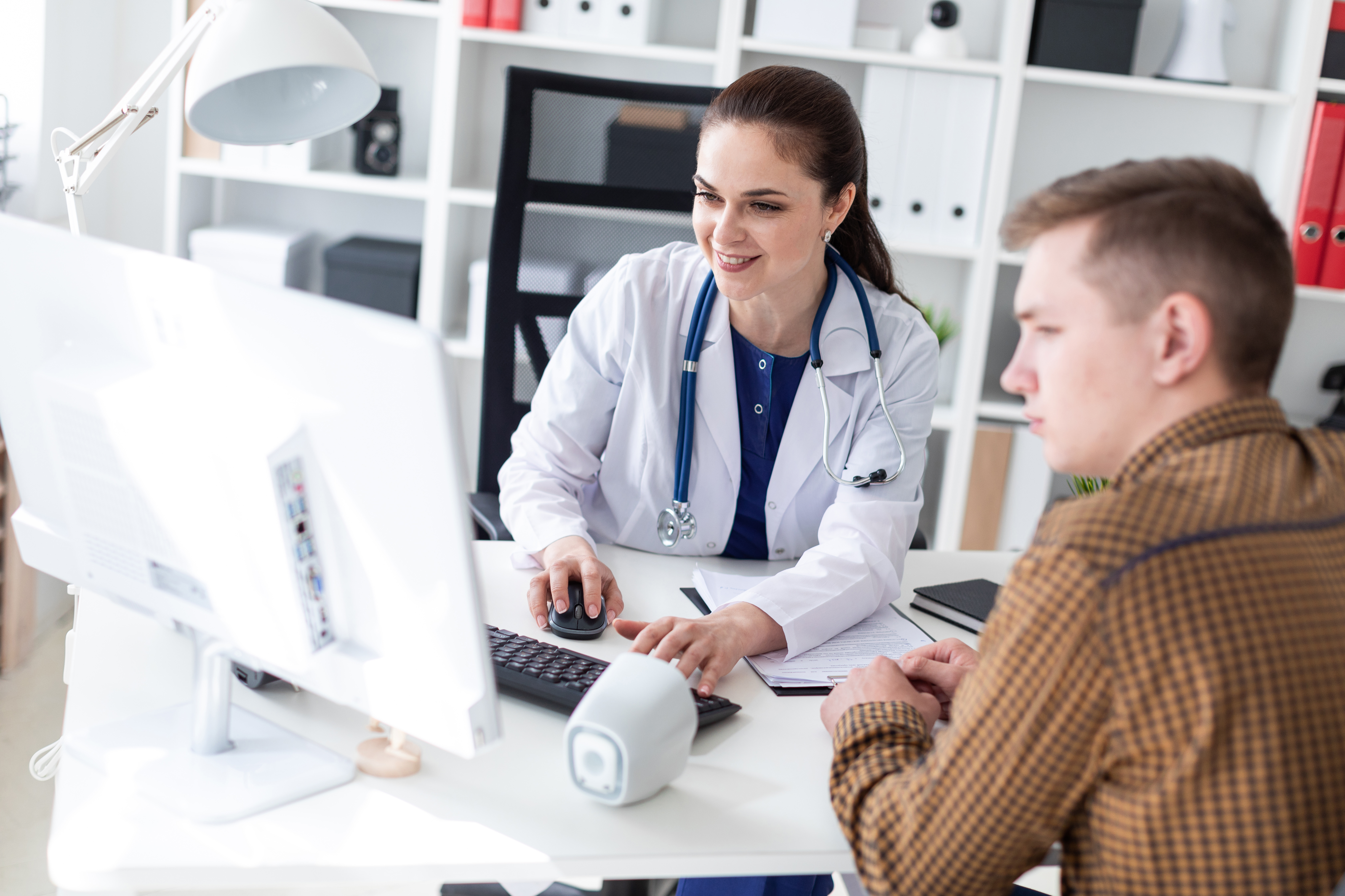 Stock photo of a female doctor in a white coat discussing information on a computer screen with a male patient in an office setting.