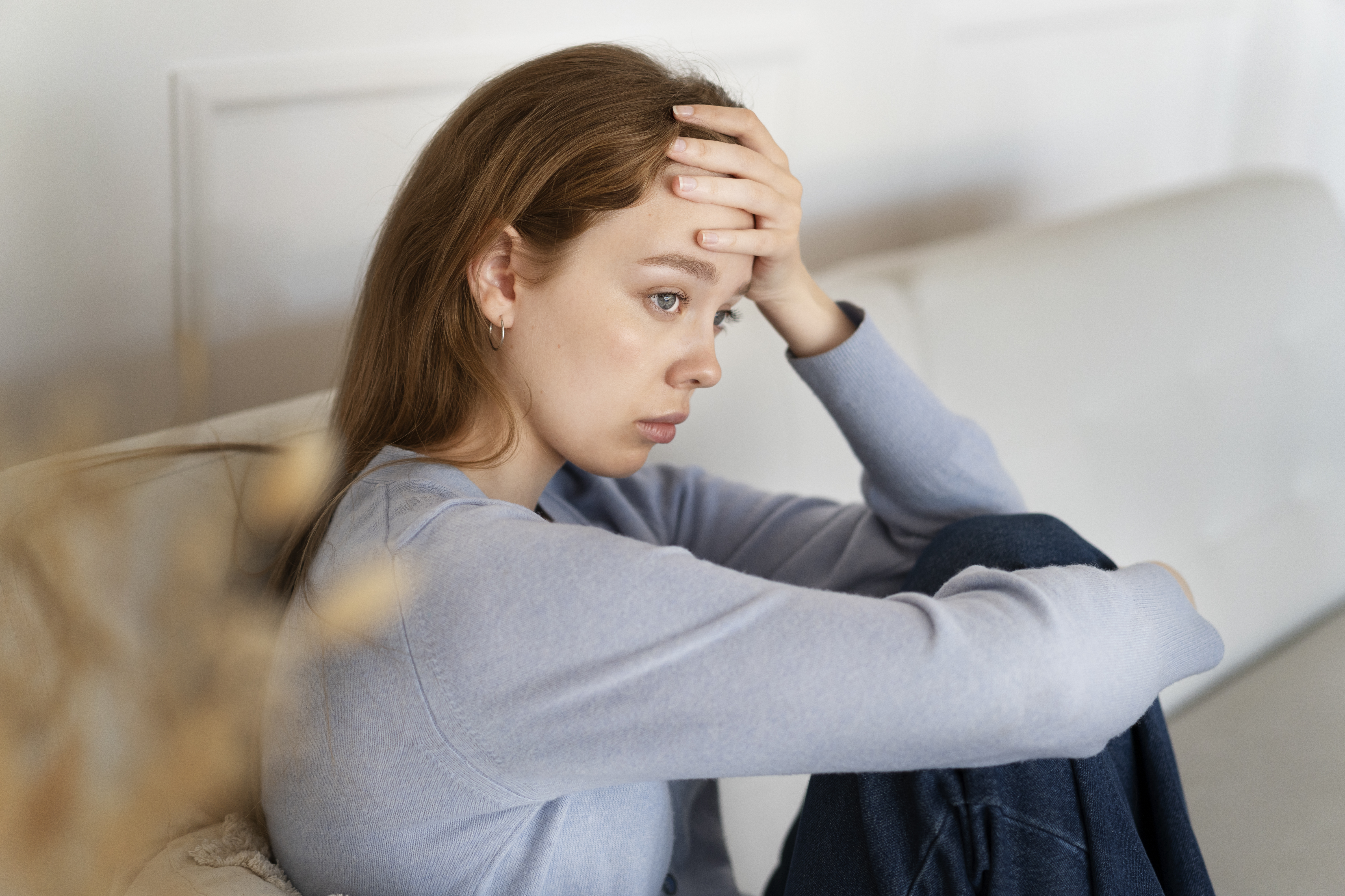 A young woman sits on a couch looking anxious and stressed, holding her forehead with her hand