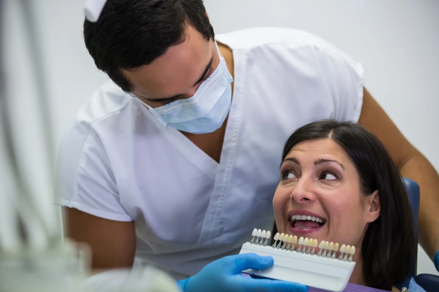 A masked dentist checks a tooth shade guide against a smiling female patient's teeth in a dental chair