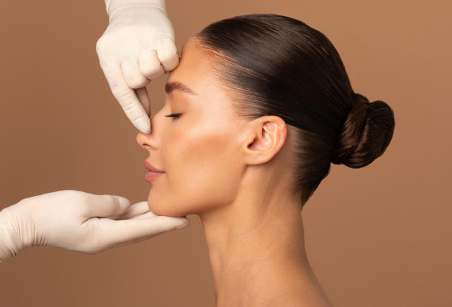 A gloved provider examines a woman's nose in profile, illustrating a rhinoplasty consultation on a blog about revision vs pri