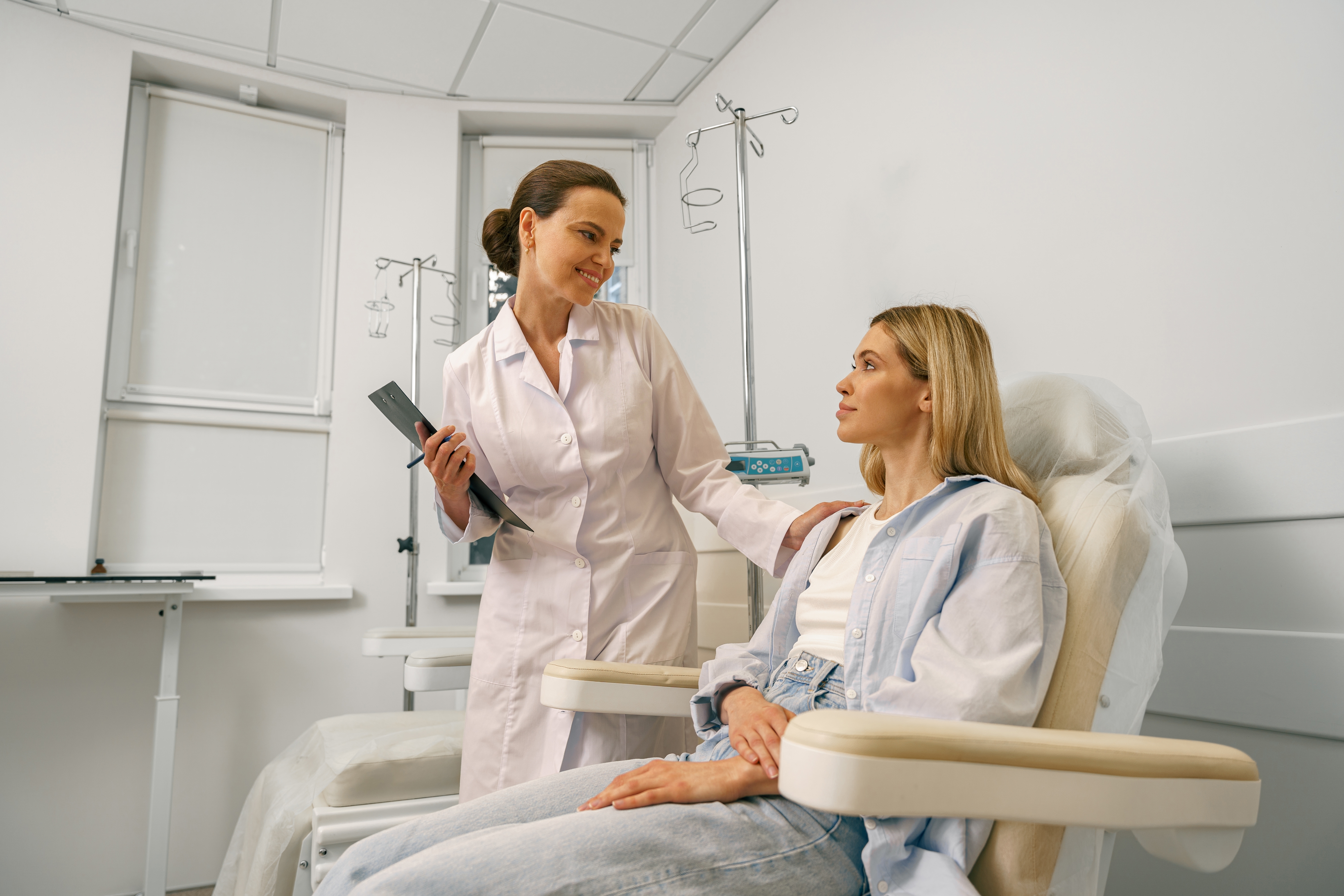 A smiling female doctor in a white coat consults with a seated patient in an infusion treatment room.