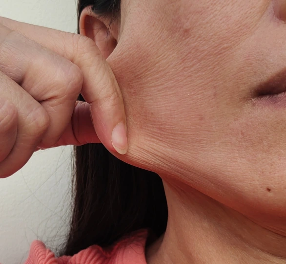 Close-up of a woman pinching the skin along her jaw, showing fine crepey lines on the cheek and neck