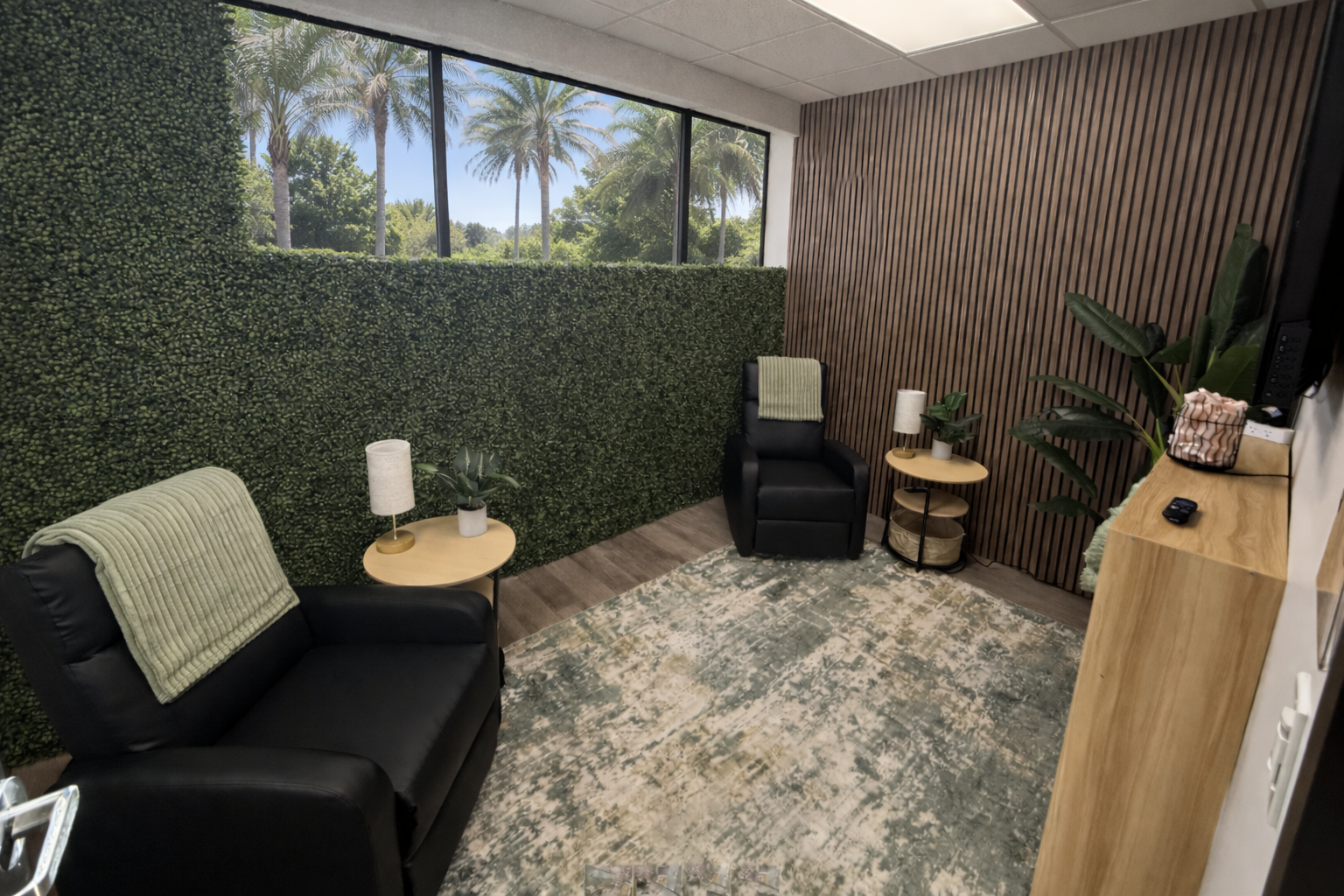 A calming treatment room with black recliner chairs, a living green wall, and a palm-tree view through large windows
