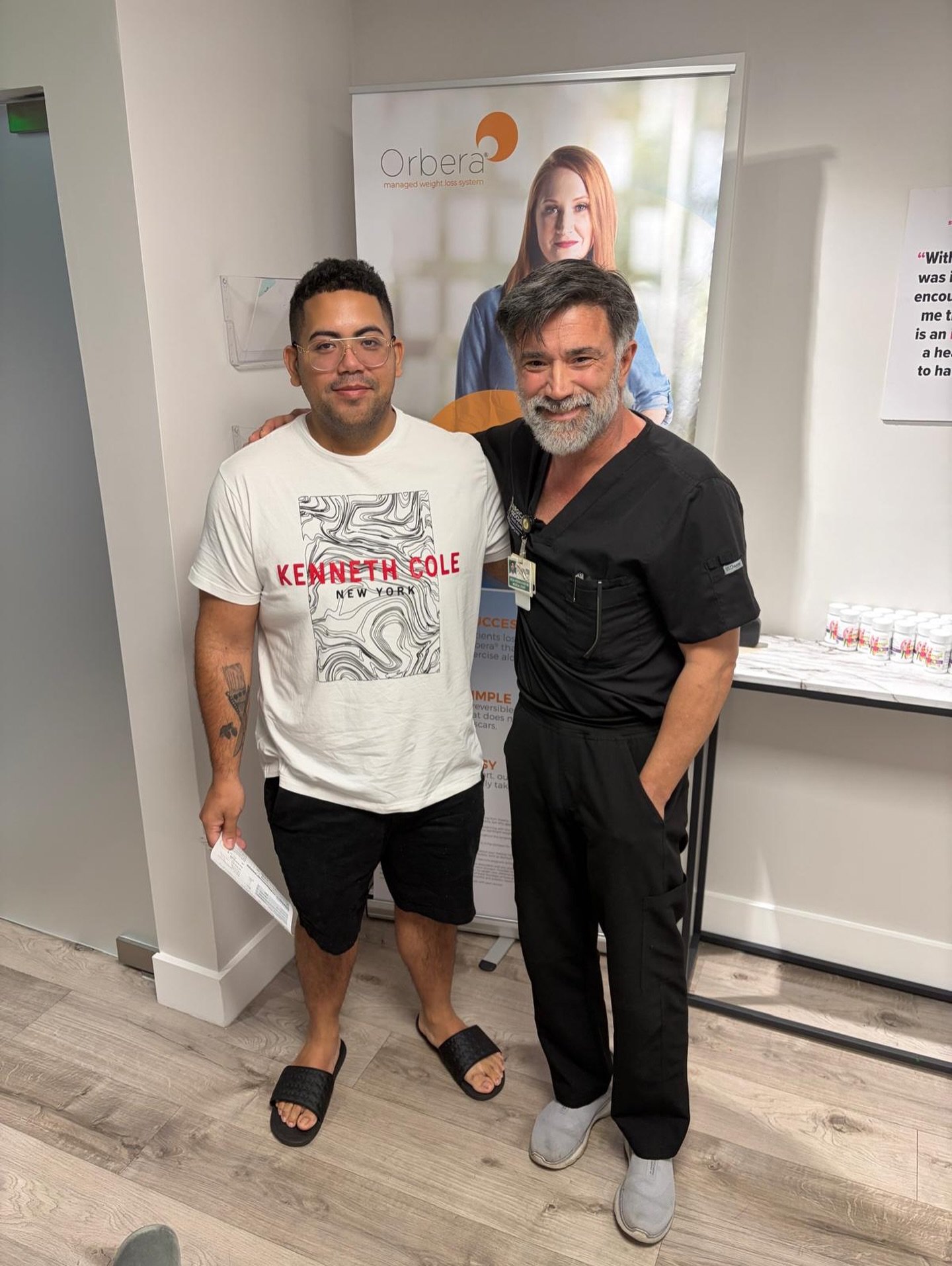 A bariatric surgery patient in a white t-shirt stands beside Dr. Keshavarzi in black scrubs in our office