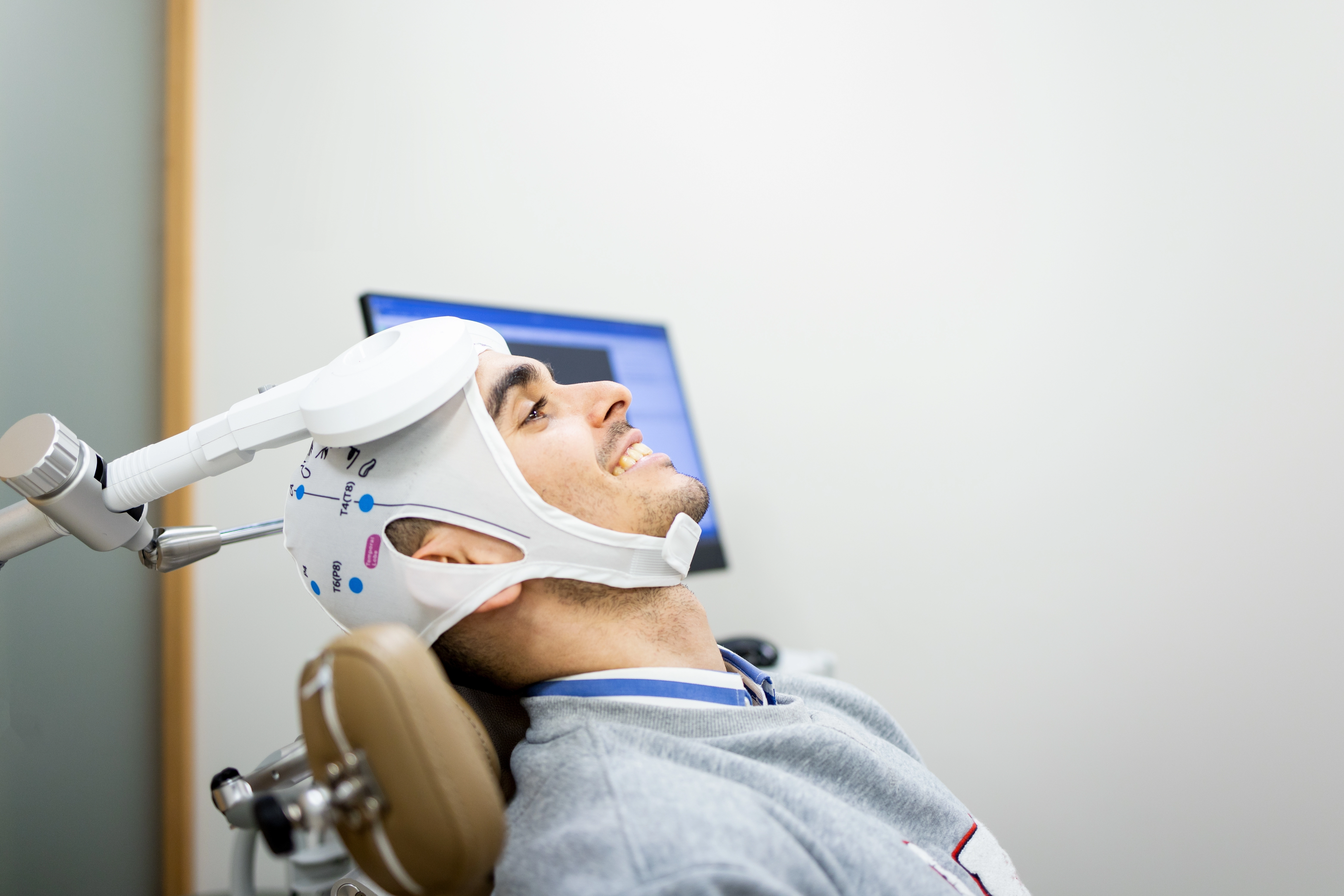 A smiling male patient reclines in a treatment chair wearing a fitted TMS coil cap during a transcranial magnetic stimulation