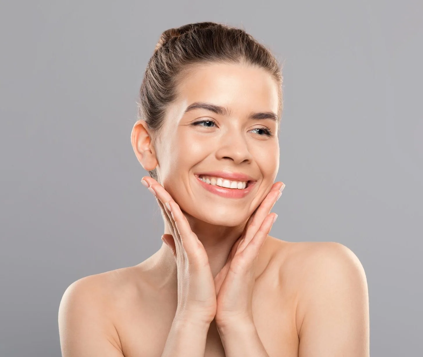 A smiling woman with clear, glowing skin touches her face against a plain gray studio background