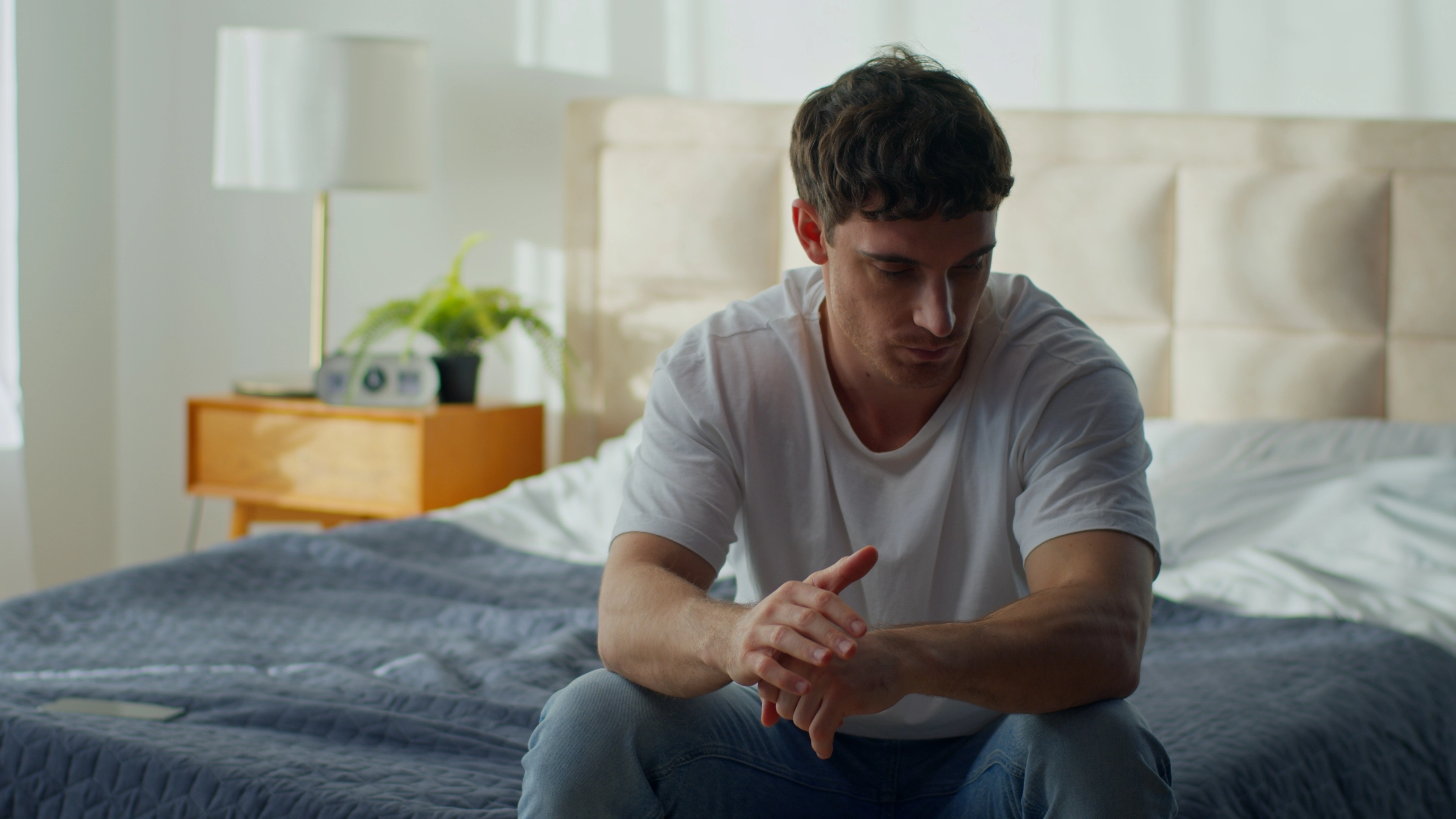 A pensive man sits on the edge of his bed with hands clasped, illustrating signs of low testosterone in men.