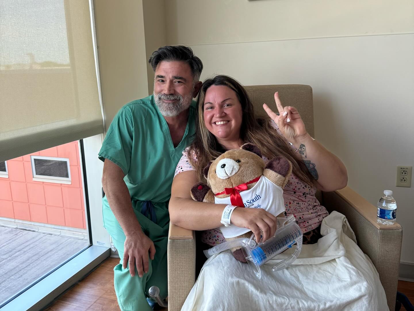 Dr. Reza Keshavarzi in green scrubs kneels beside a smiling patient holding a teddy bear in her hospital room the day after her weight loss surgery