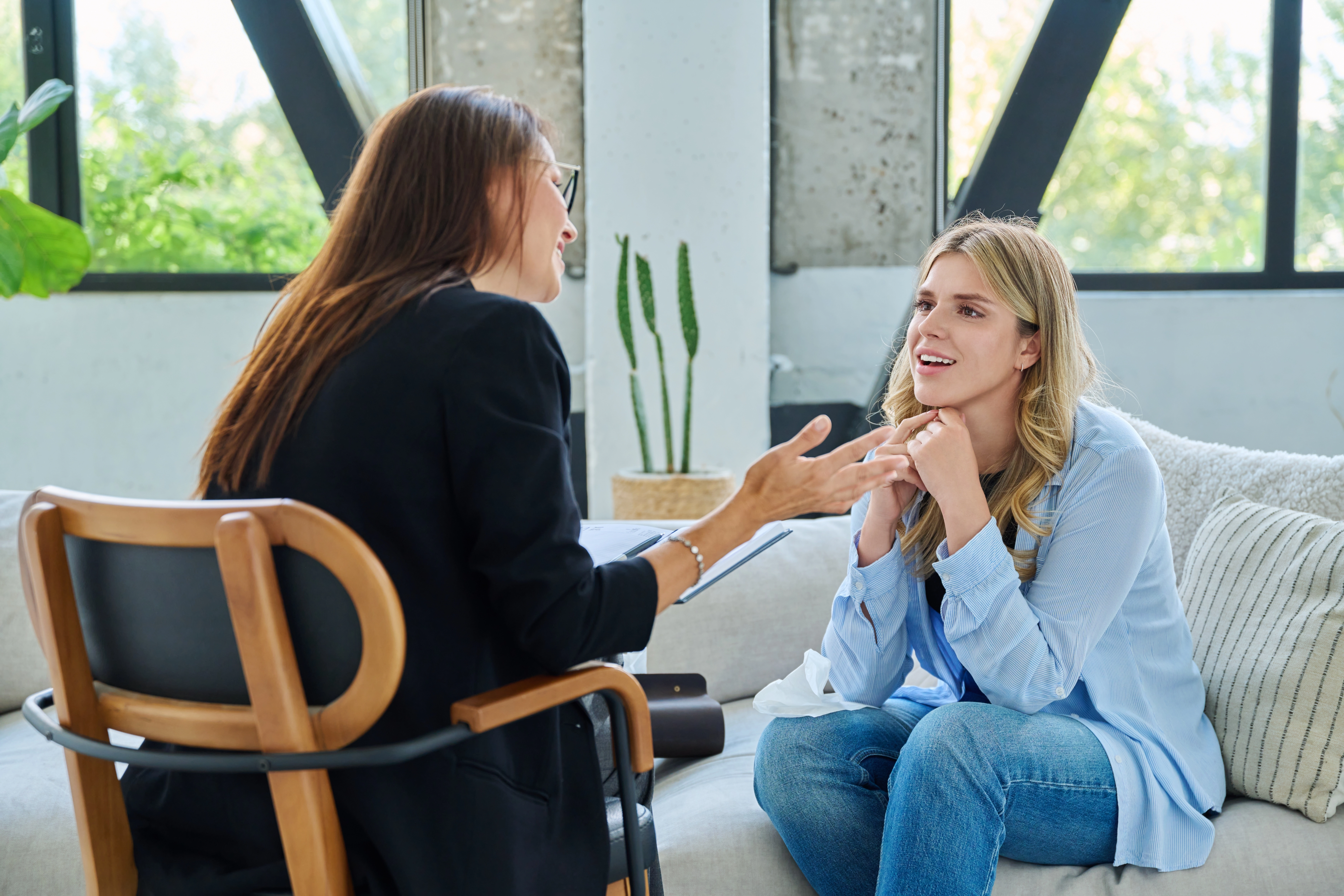 A therapist in a blazer converses with a client seated on a couch during a counseling session in a bright, plant-filled offic