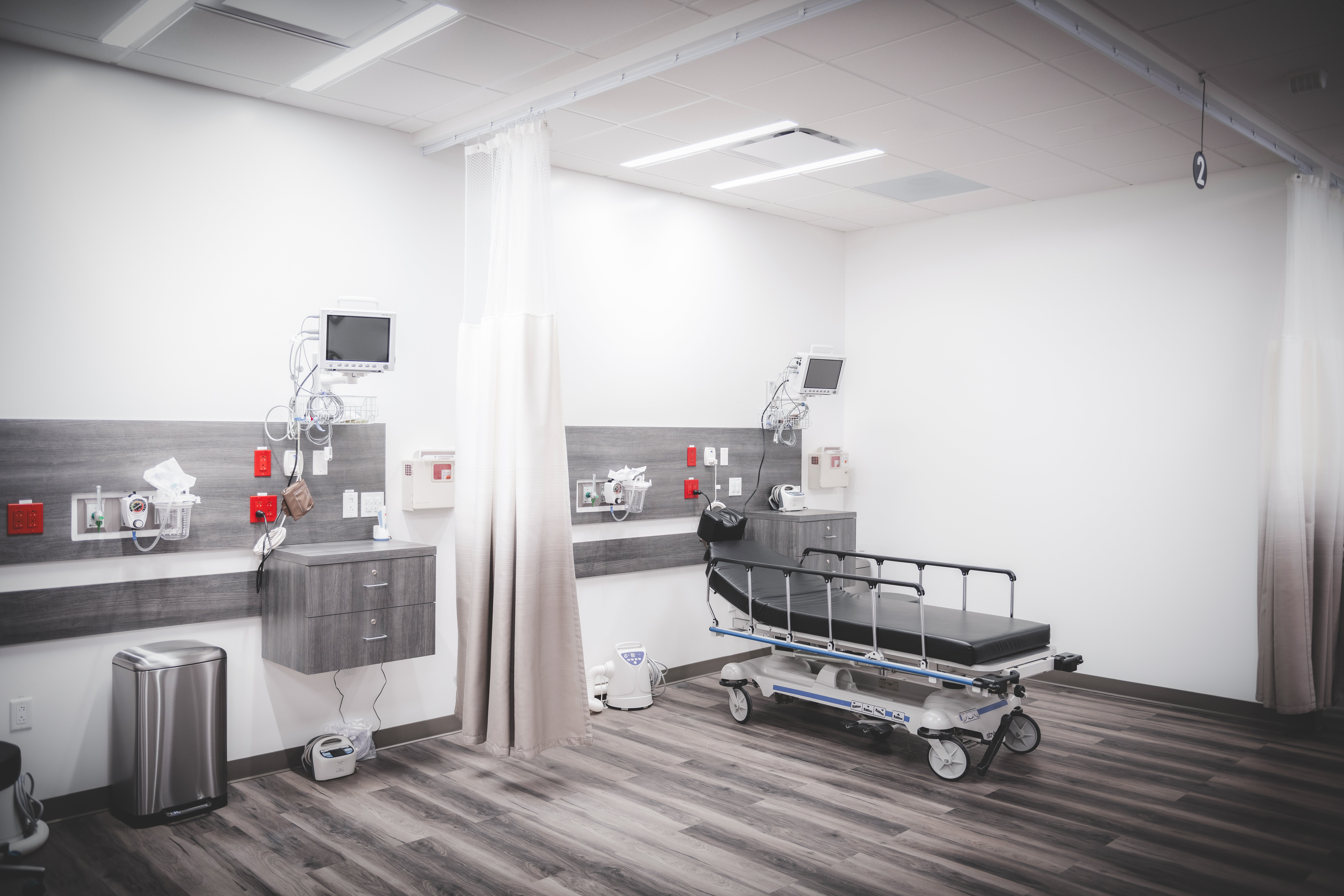 An empty clinical bay with a stretcher, privacy curtains, and wall-mounted monitors in a modern medical facility
