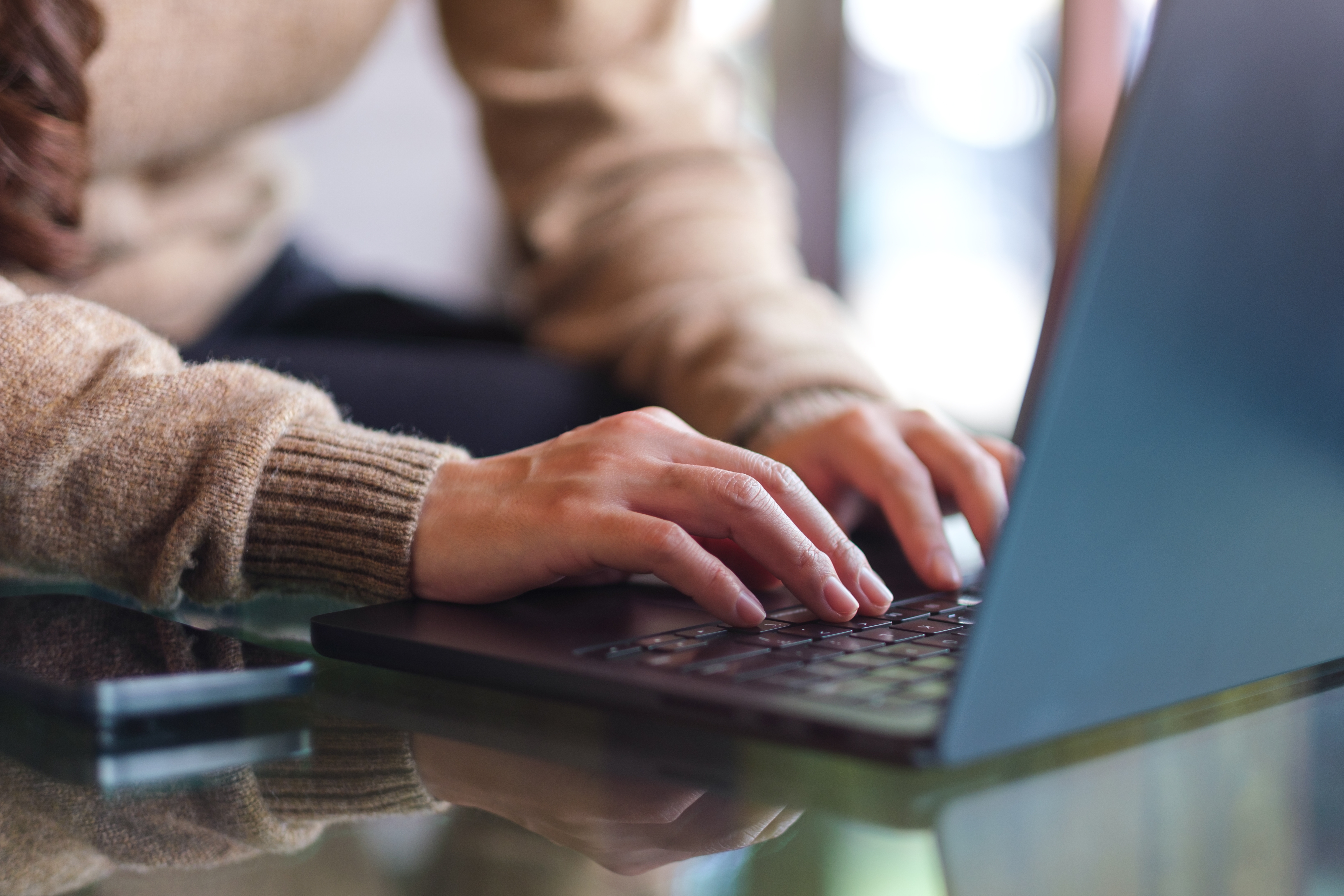 Close-up of a woman's hands typing on a laptop resting on a reflective glass table in soft natural light
