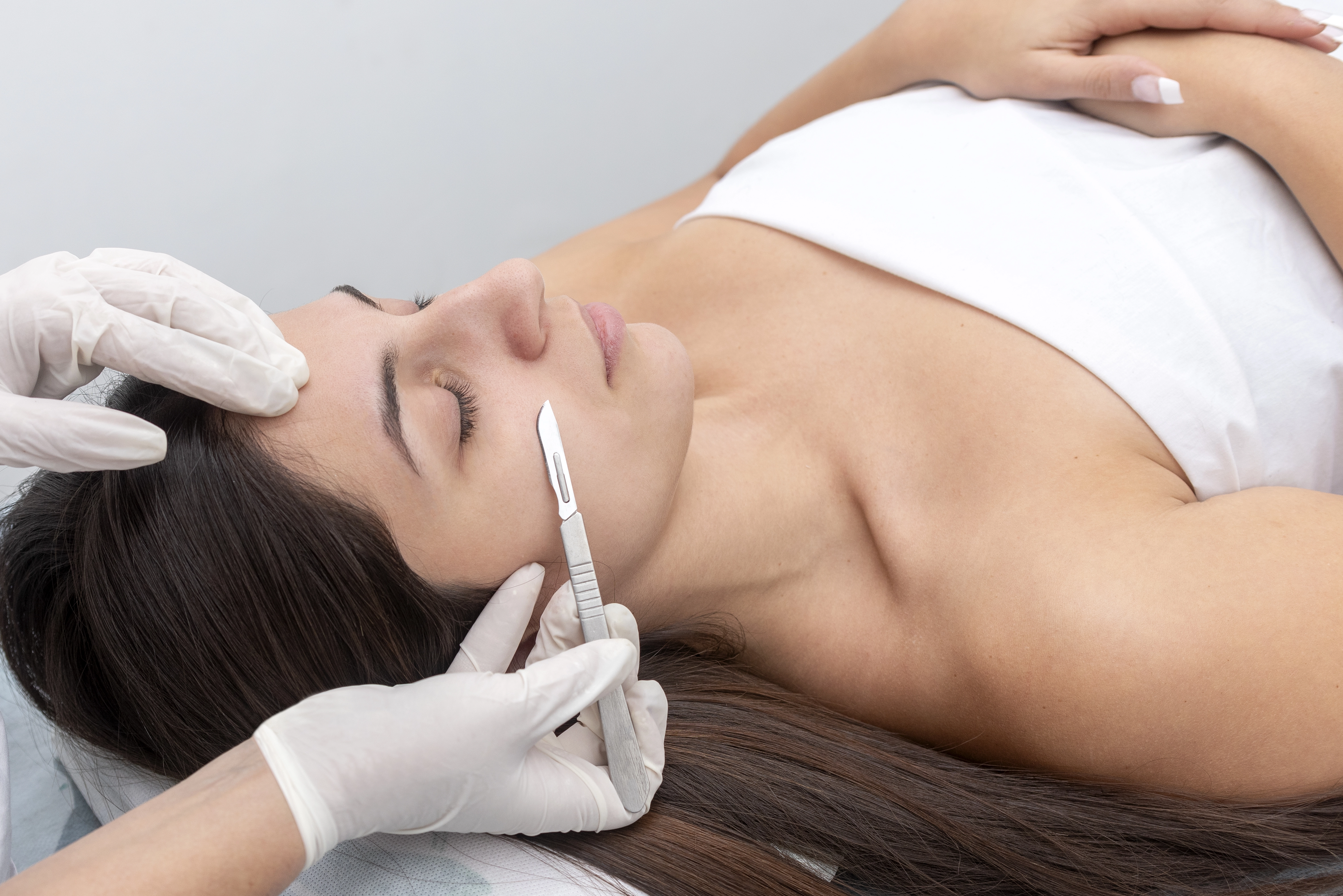 Gloved hands holding a dermaplaning blade at a shallow angle against a relaxed woman's cheek