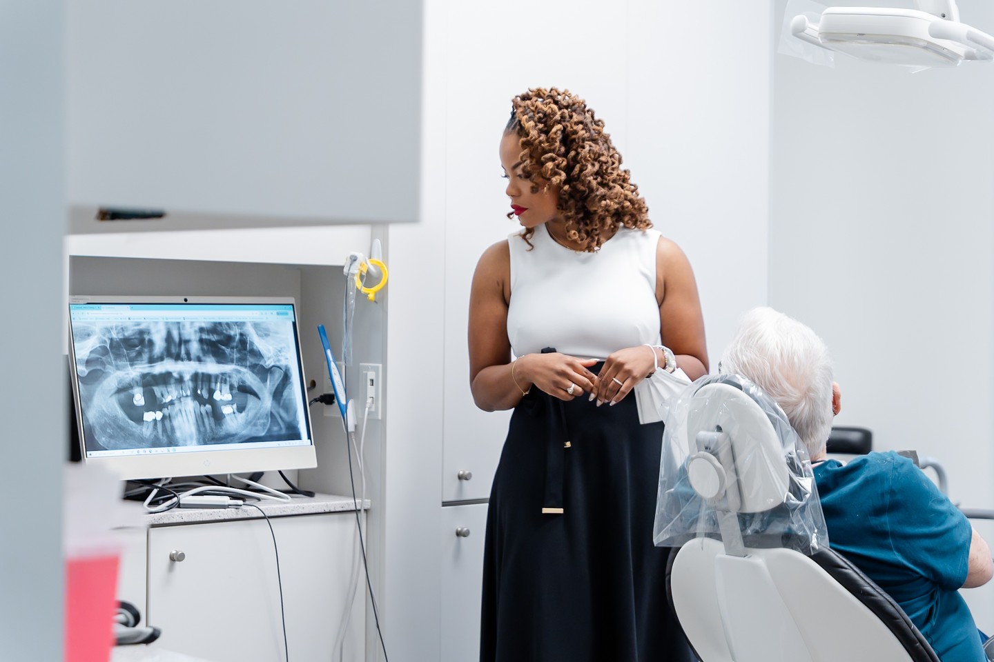 A dental staff member reviews a dental x-ray on a monitor while a patient sits in the exam chair beside her