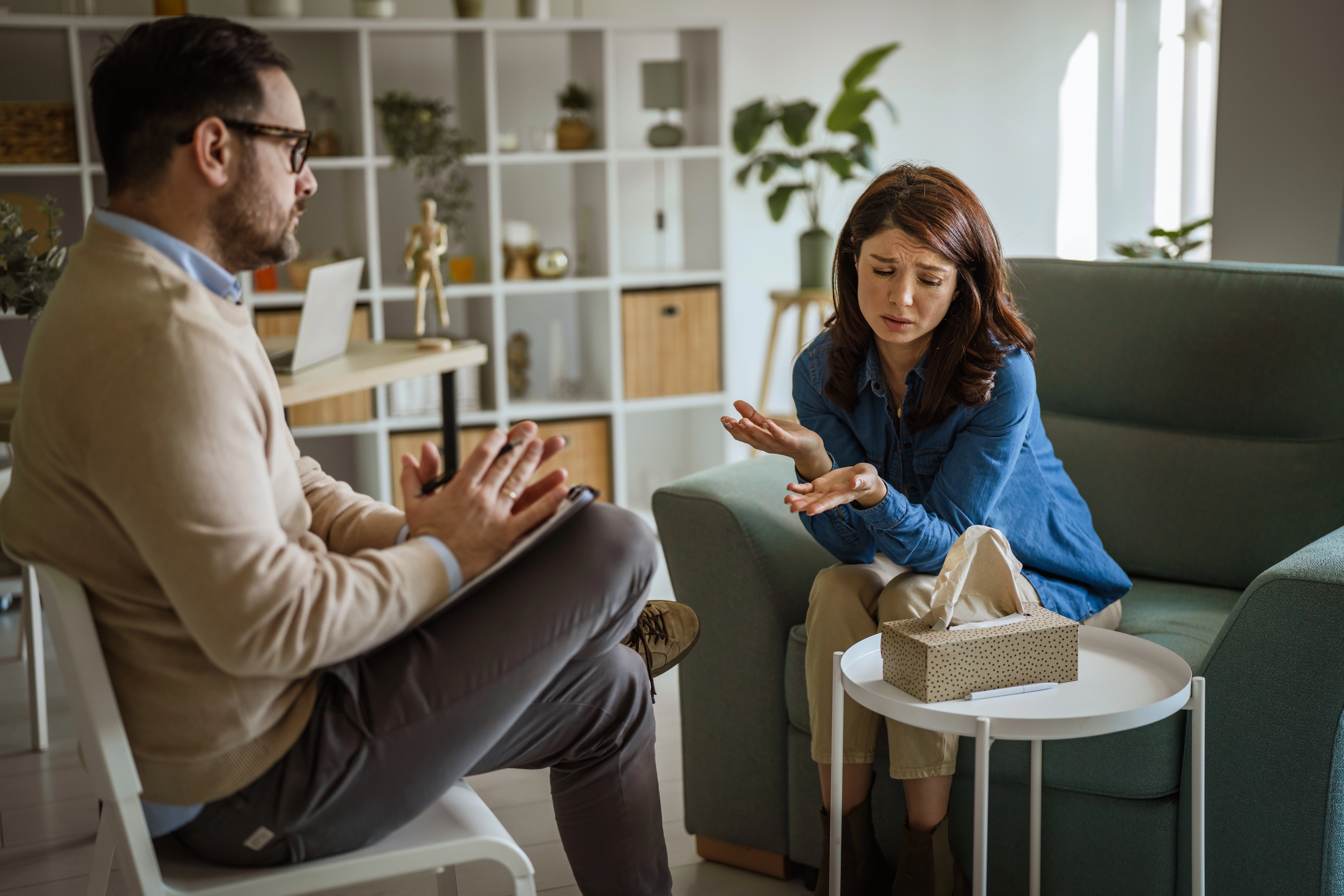 A male therapist listens attentively while a distressed female client gestures during a counseling session in a softly lit of