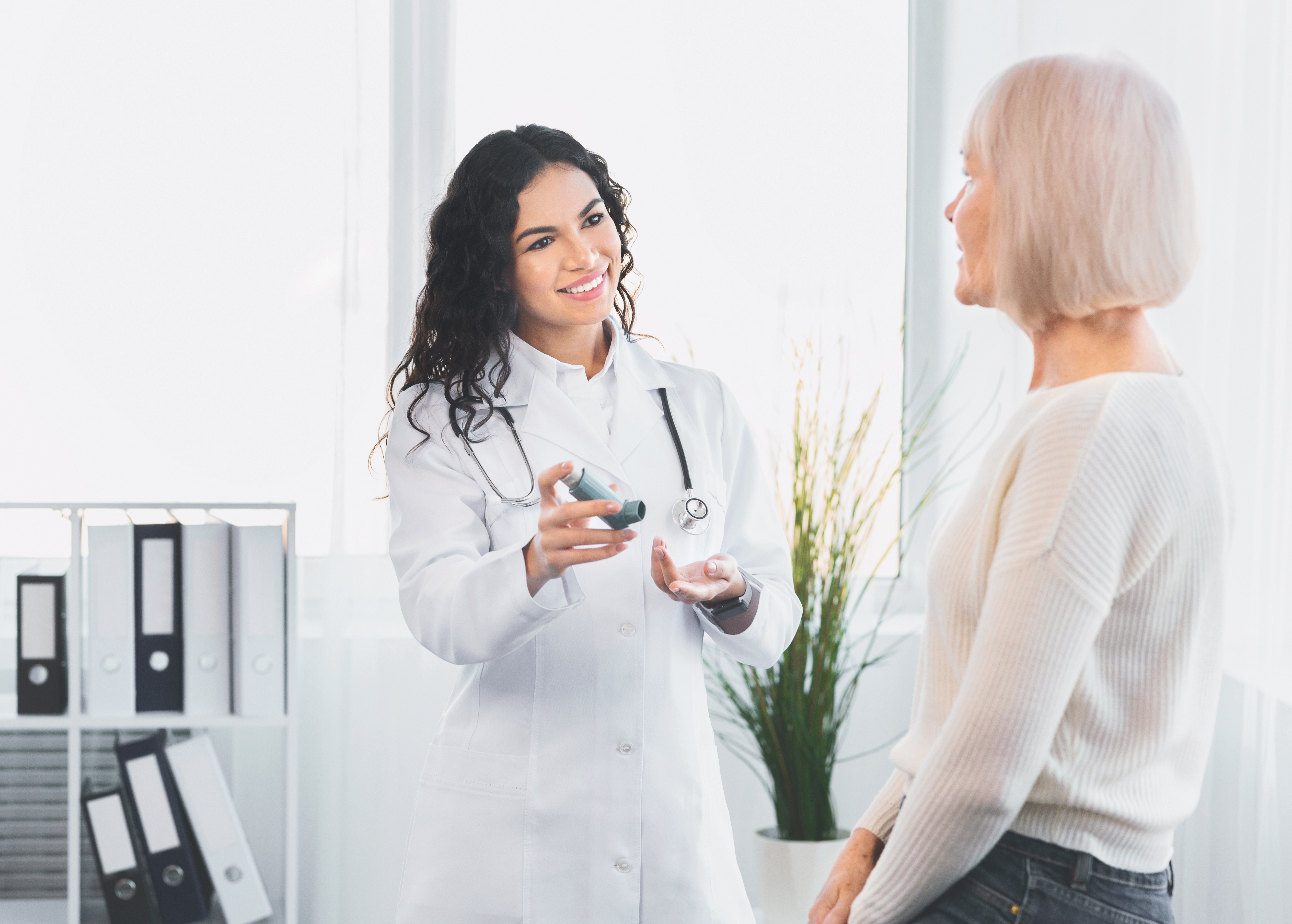 A female doctor in a white coat explains use of an inhaler to an older female patient during a consultation about asthma or COPD.