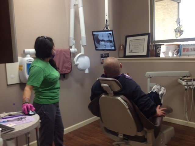 A dental assistant in a green shirt reviews a dental x-ray on a wall monitor with a seated patient during a treatment visit