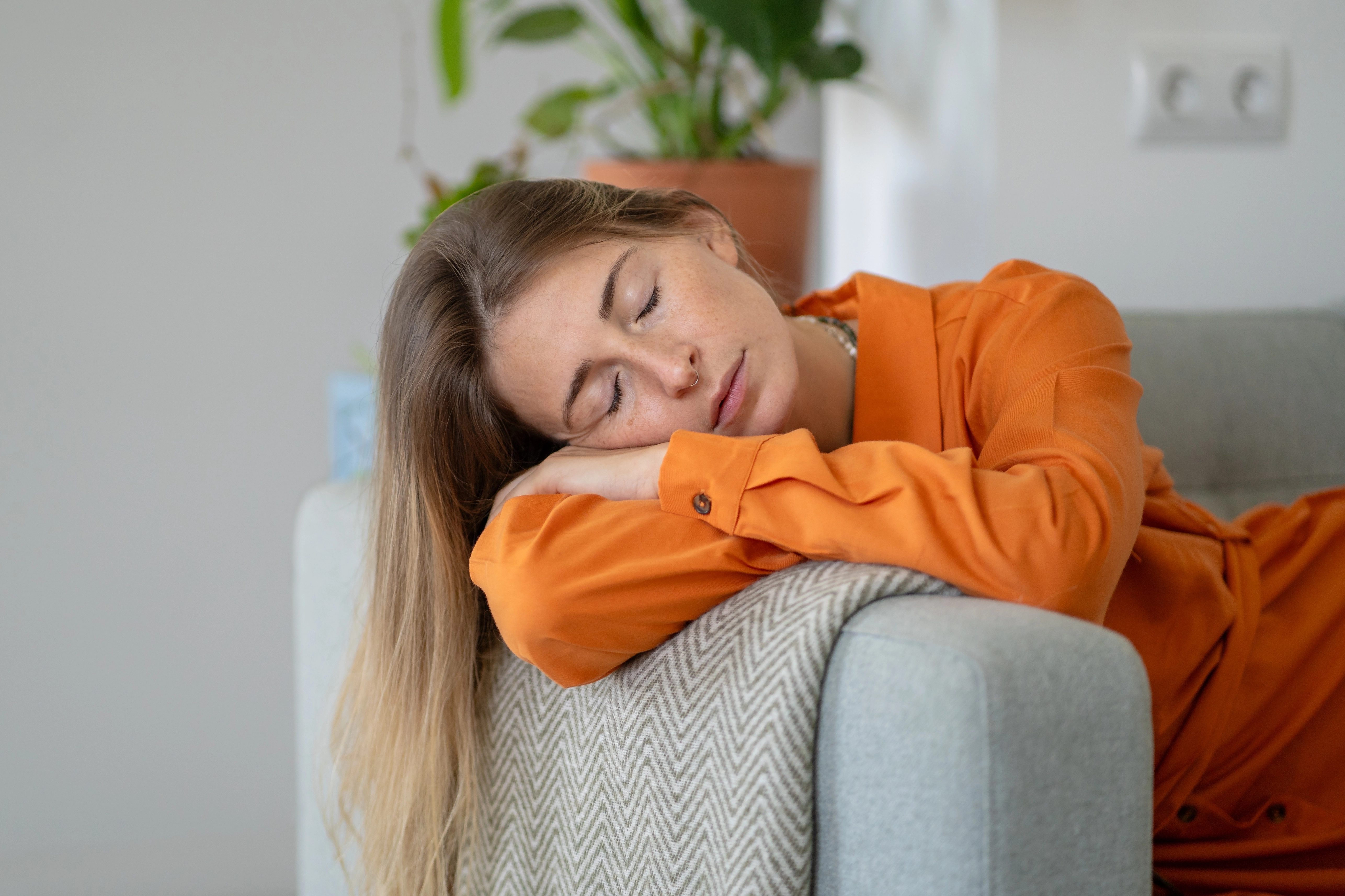A woman in an orange shirt rests her head on a couch pillow with her eyes closed, illustrating fatigue at home