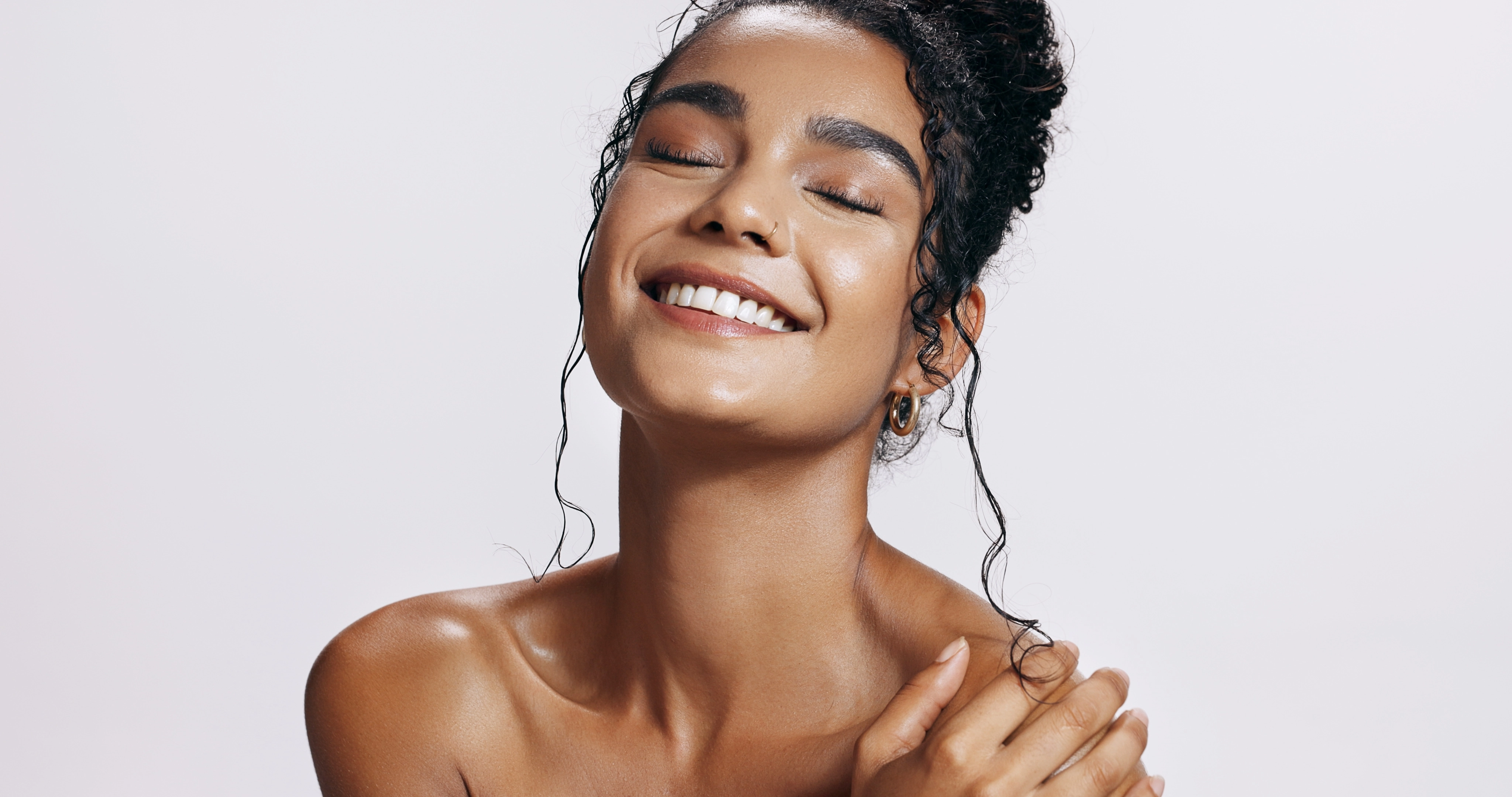 A smiling woman with glowing skin and wet hair tied up poses against a plain light background, showcasing radiant skin