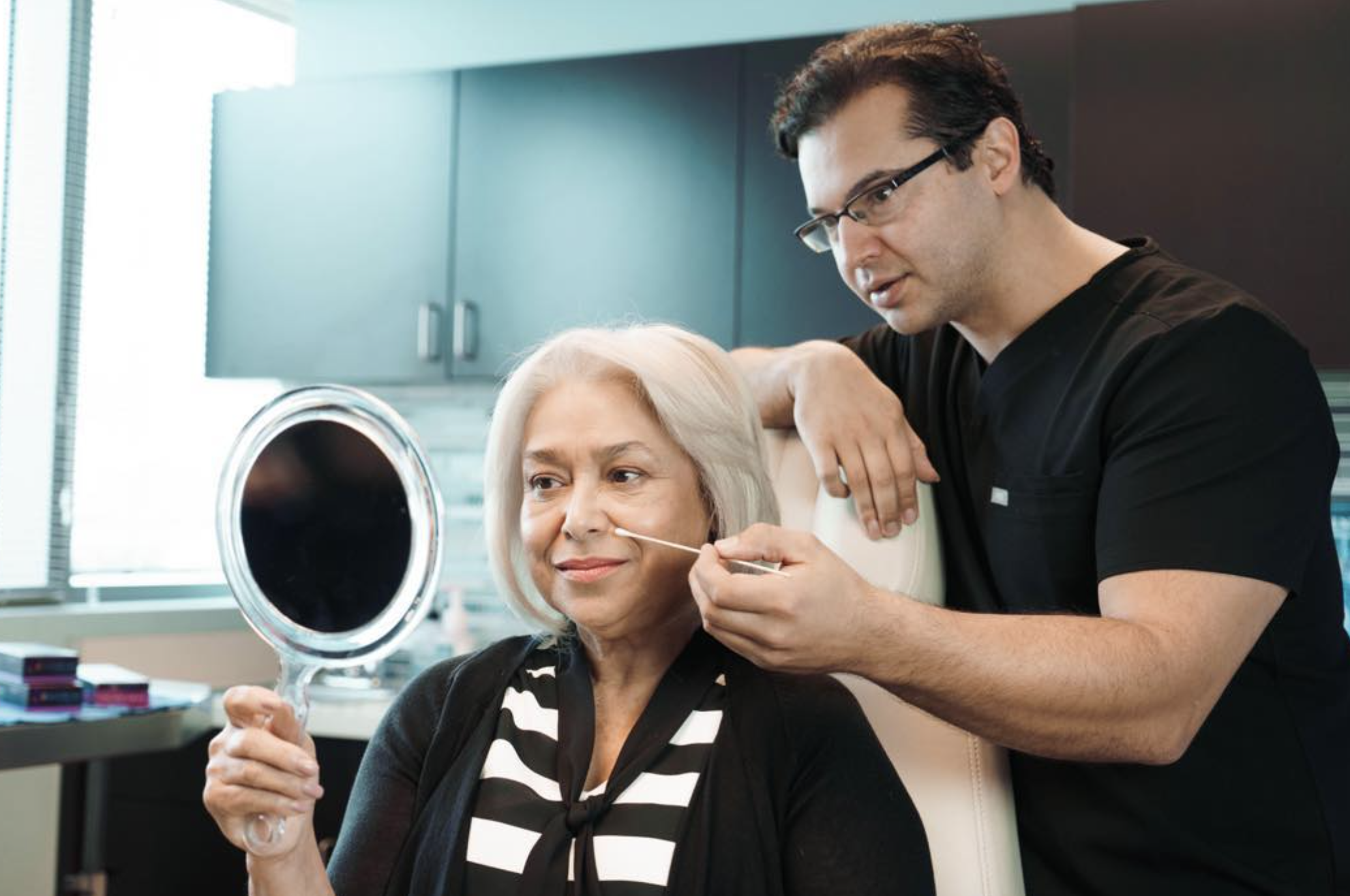 A male provider examines an older woman's face with a cotton swab while she looks in a hand mirror in a clinical office