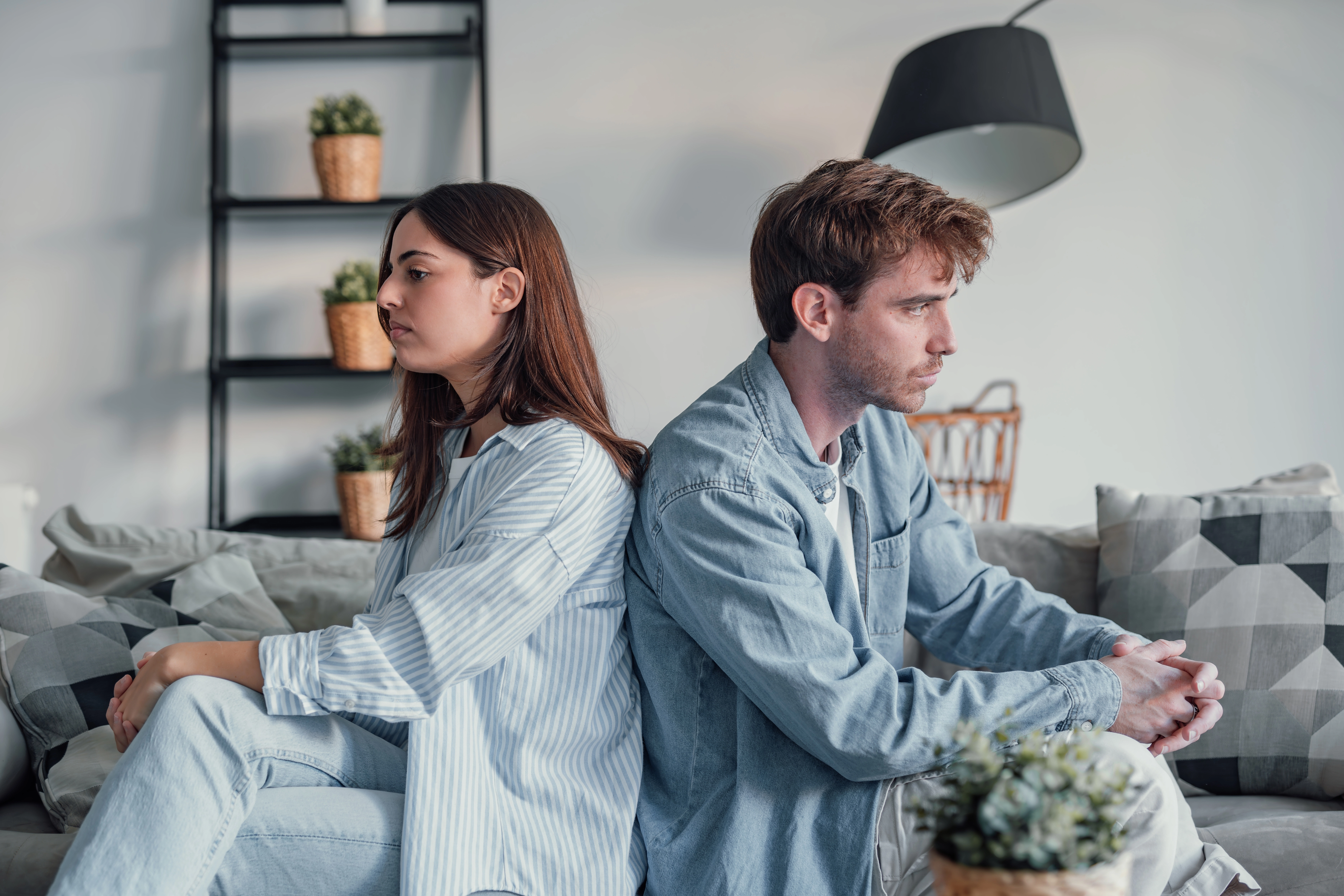 A couple sits back-to-back on a sofa with tense, distant expressions, illustrating relationship conflict or communication str
