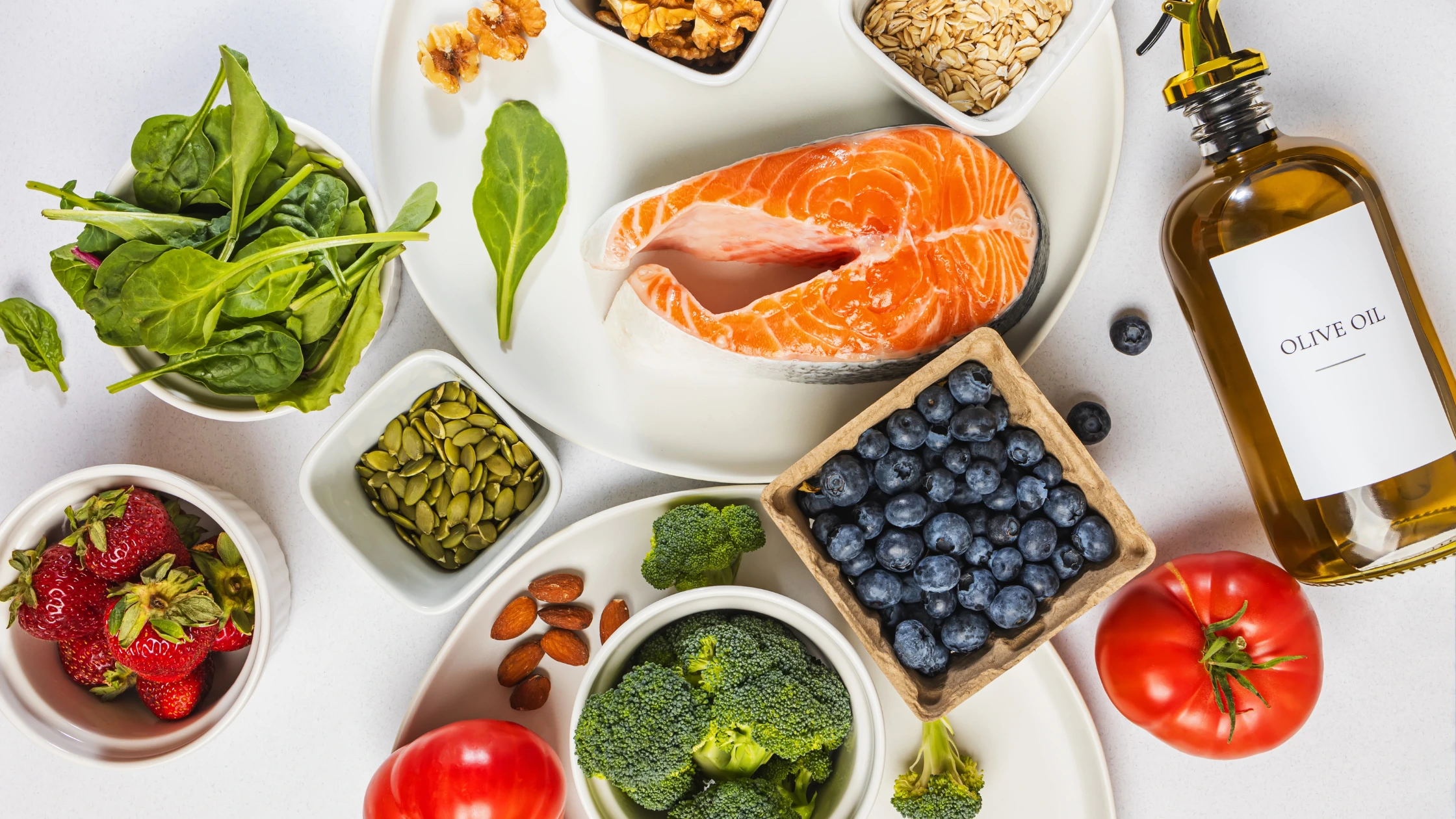 An overhead flat-lay of nutritious whole foods including salmon, vegetables, berries, nuts, and olive oil arranged on a white