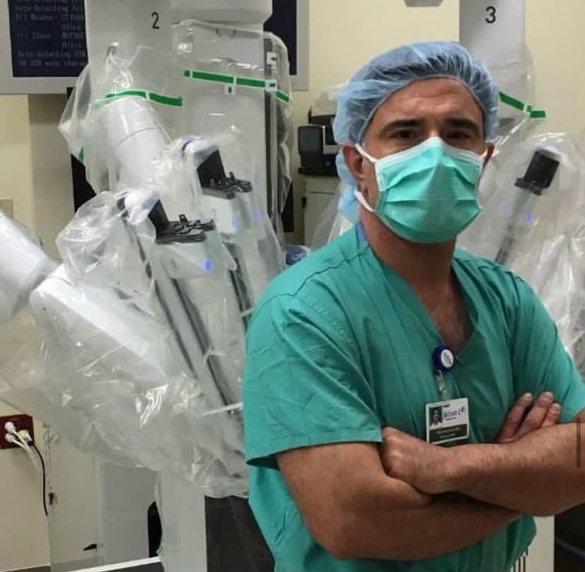 Dr. Reza Keshavarzi in green scrubs and a surgical mask, arms crossed, beside a surgical robot in the operating room