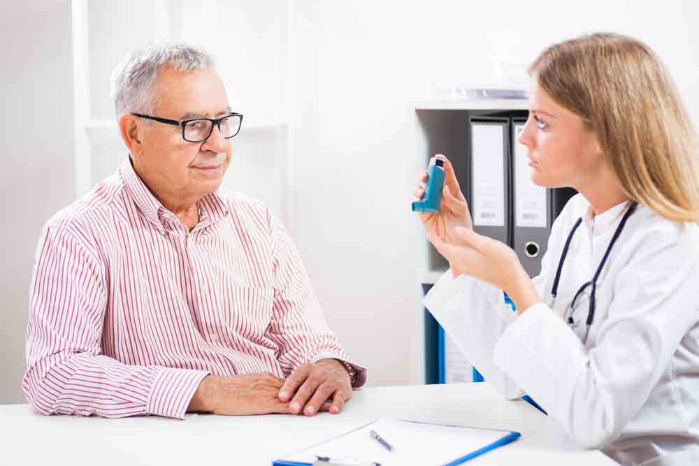 A female doctor explains the use of an asthma inhaler to an elderly male patient during a consultation.