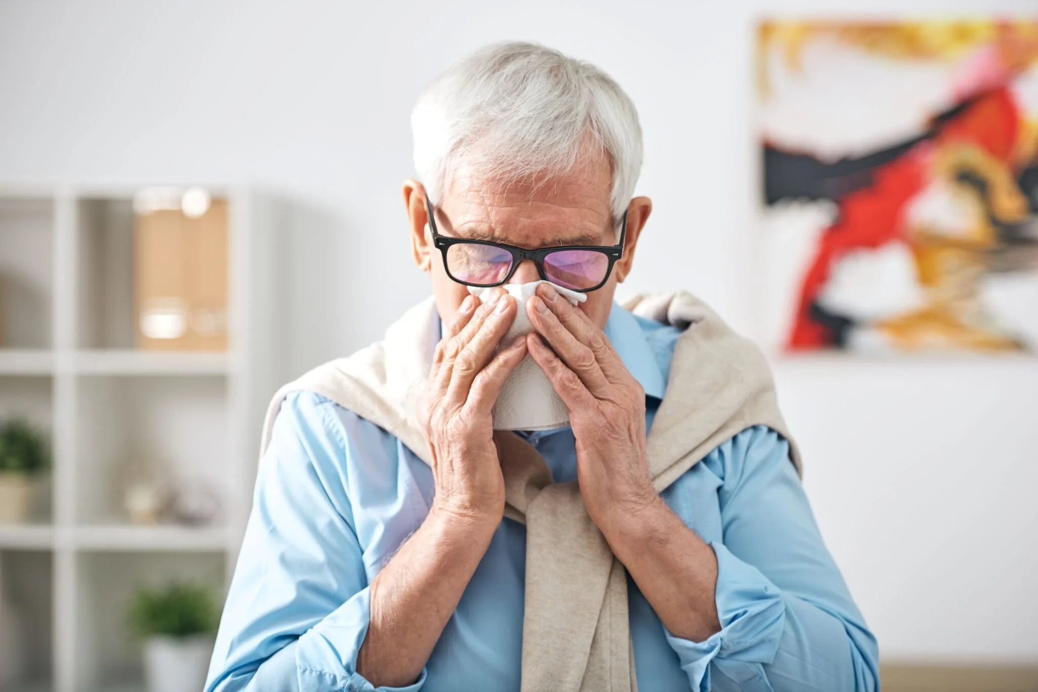 Older man in glasses blowing his nose with a tissue in a softly lit living room