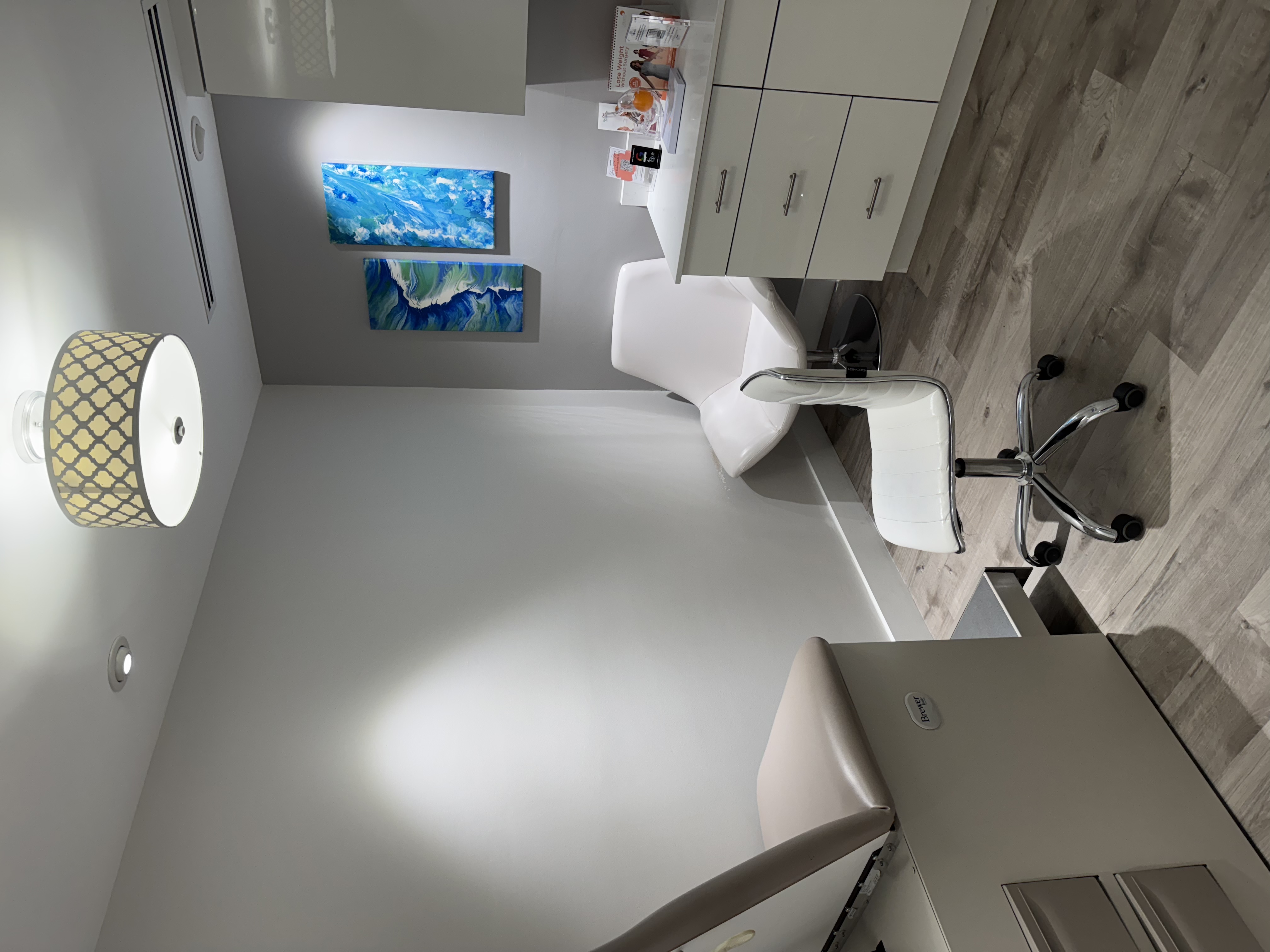 Exam room at our South Miami office with an exam table, white rolling chair, cabinetry and ocean artwork