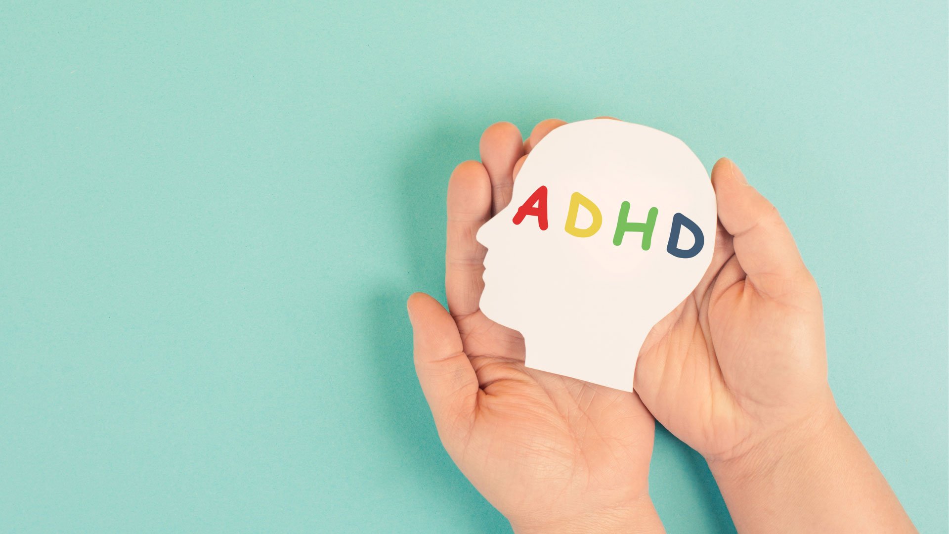 Hands hold a paper cutout of a head with the word ADHD colorfully lettered across it against a teal background