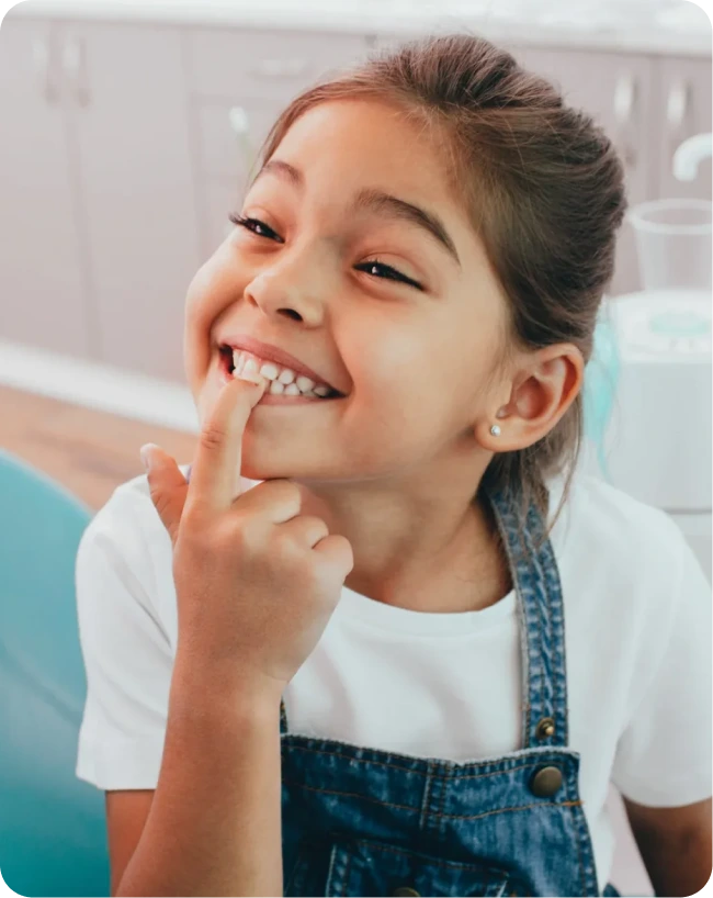 A smiling young girl points to her teeth while sitting in a dental chair