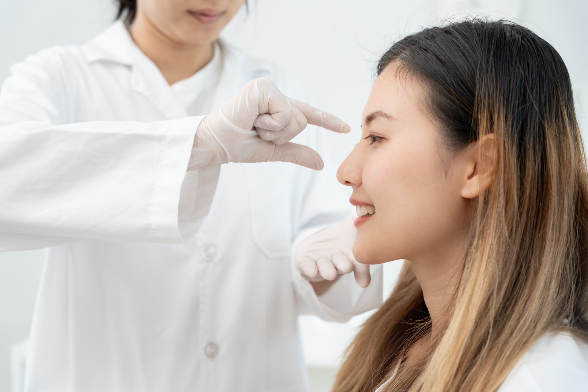 A gloved provider in a white coat examines a smiling woman's nose during a rhinoplasty consultation