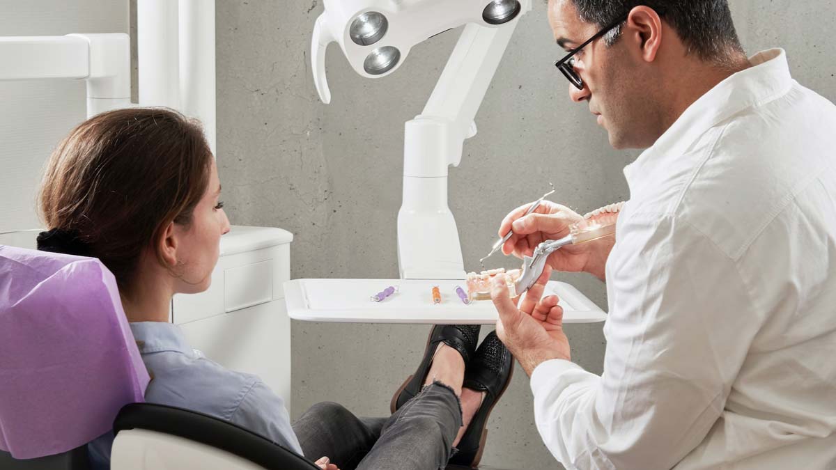 A dentist explains a dental crown using a tooth model to a seated patient in a dental exam chair