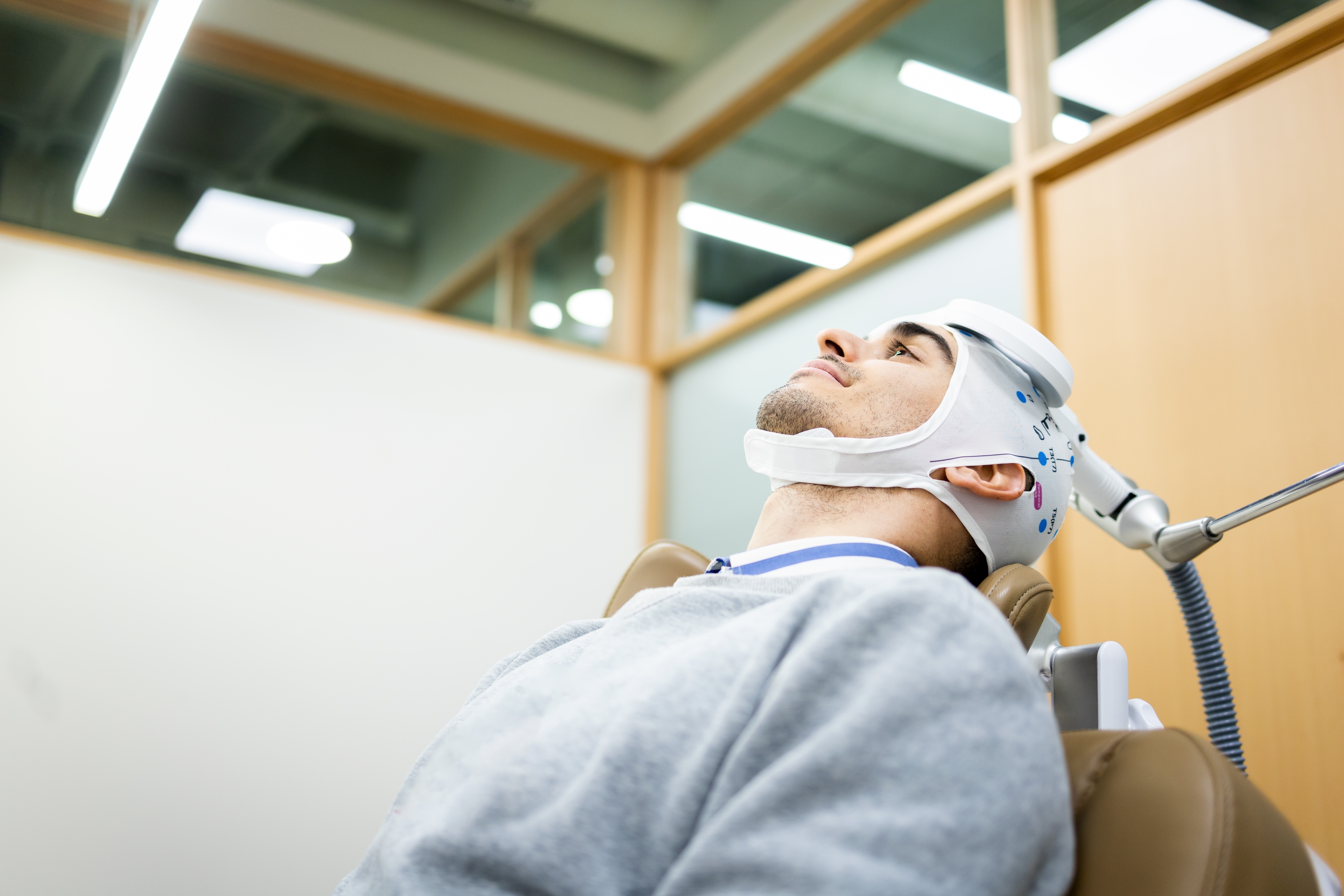 A man reclines in a treatment chair while wearing a TMS coil headpiece during a transcranial magnetic stimulation therapy ses