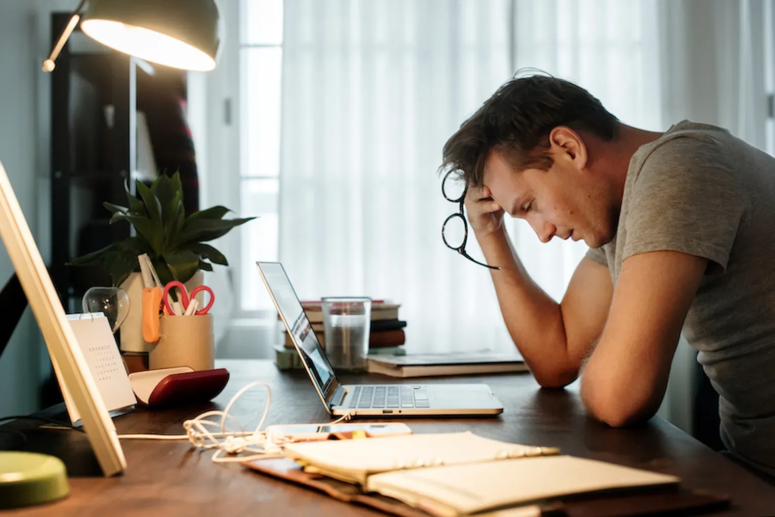 A stressed man sits at a home desk holding his glasses to his forehead while looking down at his laptop, illustrating anxiety