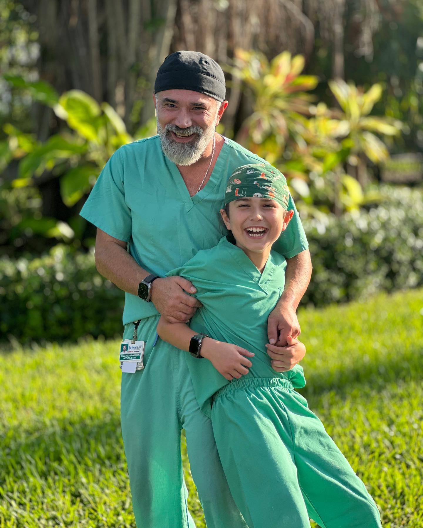 Dr. Reza Keshavarzi smiling outdoors in green surgical scrubs with his young son