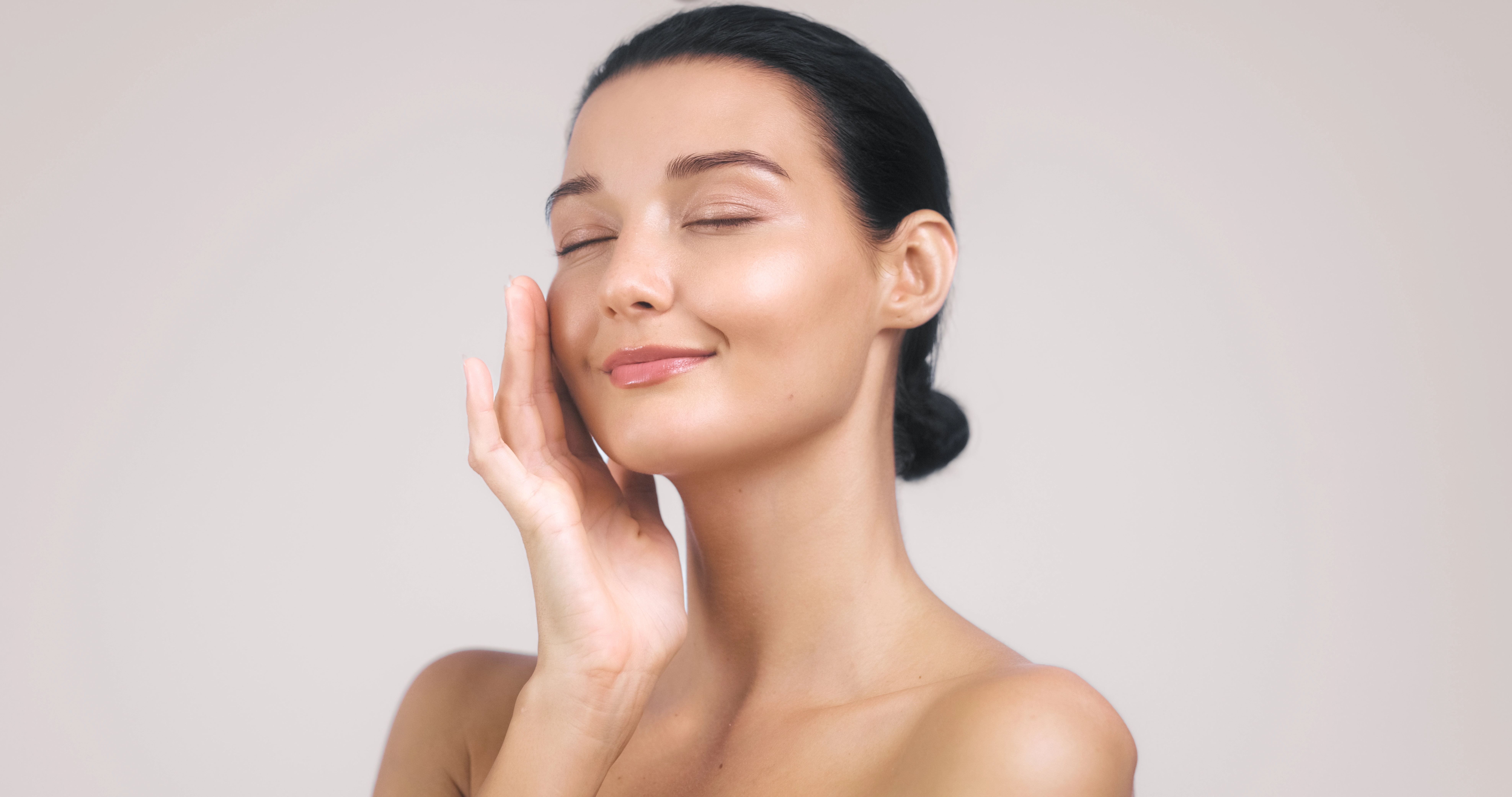 A woman with closed eyes gently touches her smooth, glowing cheek against a soft neutral background