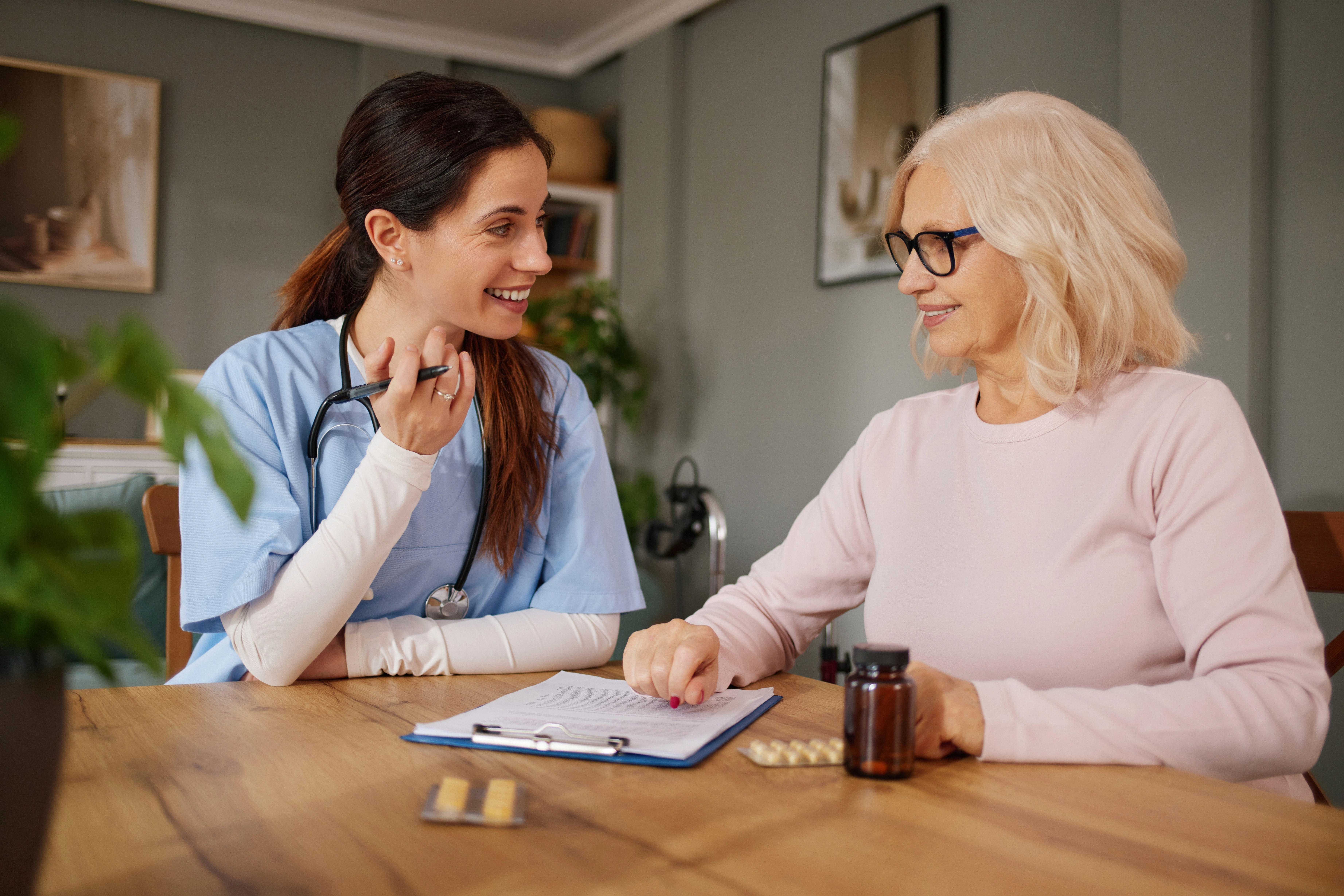 A smiling nurse practitioner reviews a medication management plan with an older female patient at a table with pill bottles n