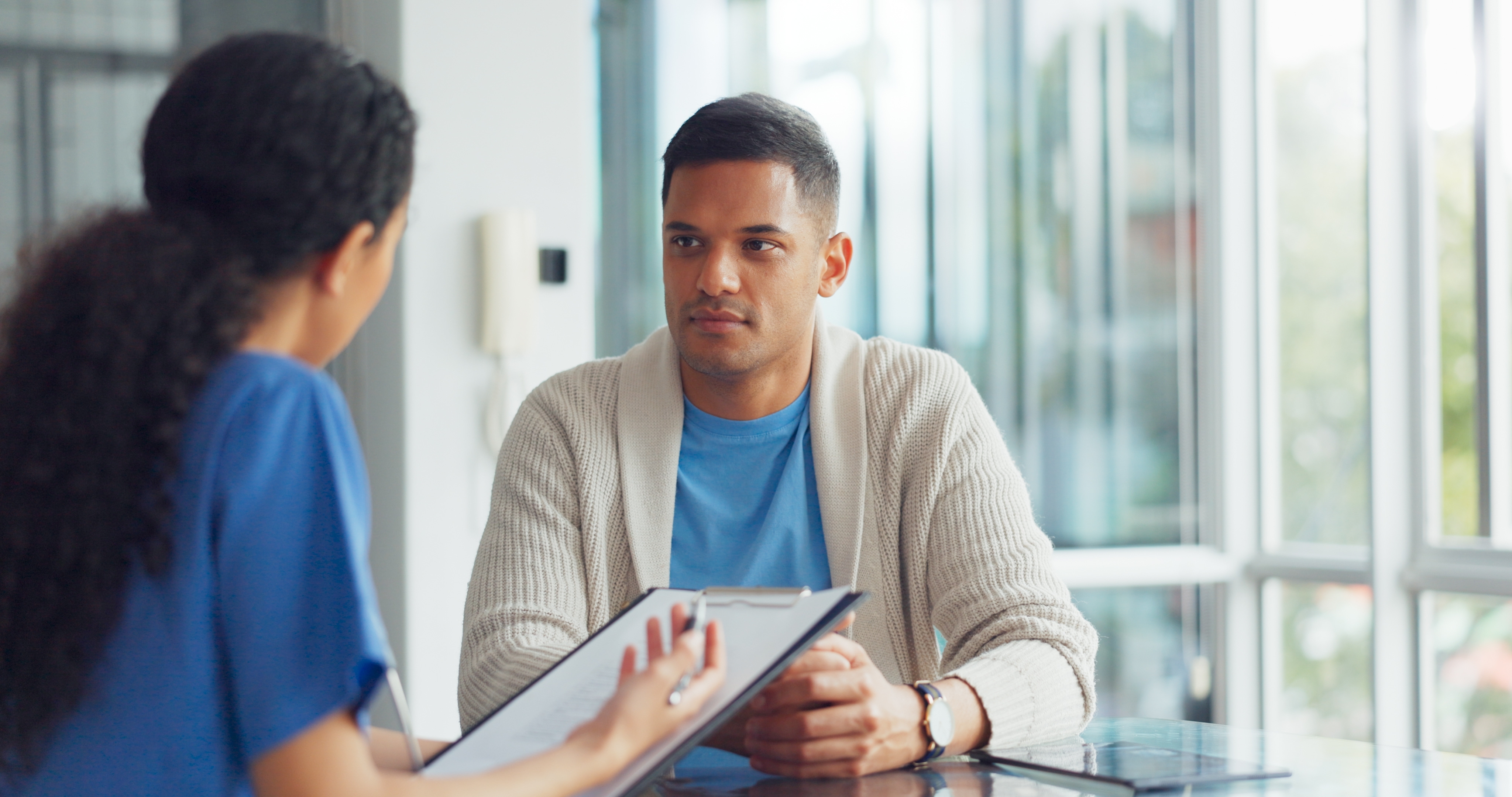 A male patient listens attentively during a consultation with a provider holding a clipboard in a bright office