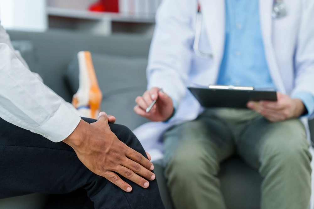 Image: A patient seated in our treatment room, holding a hand to an aching knee while discussing joint pain with our provider.