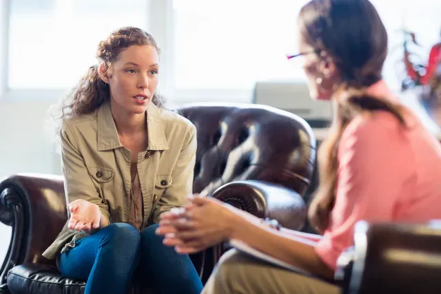 Image: A calm, warmly lit room with two comfortable chairs facing each other, suggesting a supportive conversation about trauma and recovery.