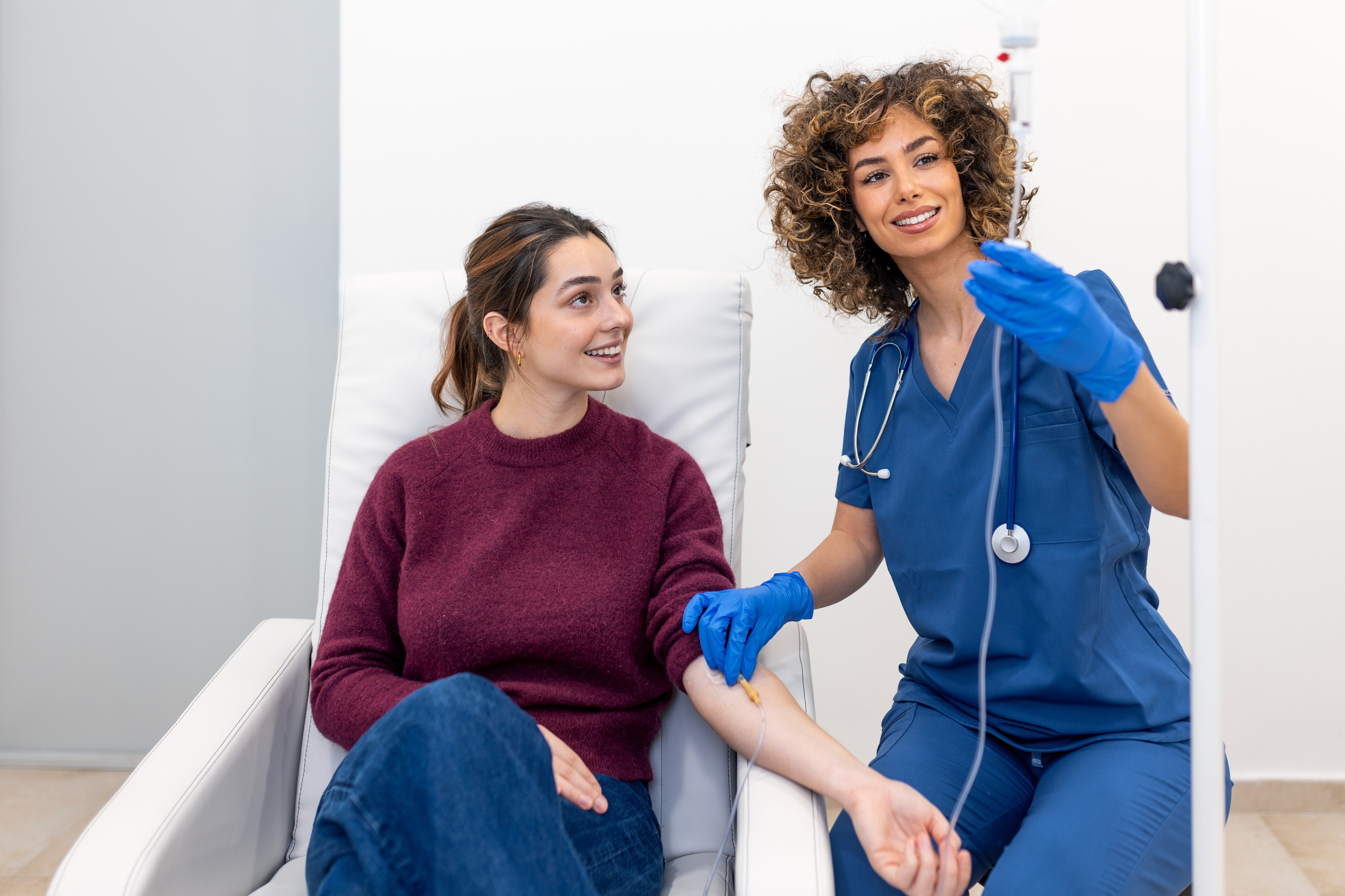 A smiling nurse in blue scrubs administers IV therapy to a relaxed female patient seated in a treatment chair.